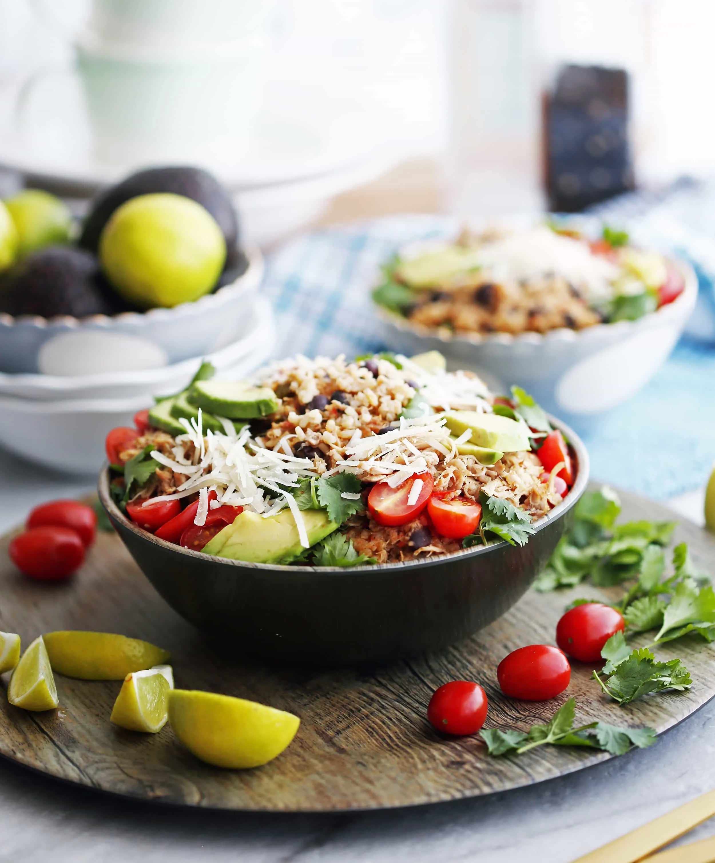 Side view of a bowl of Instant Pot Chicken and Rice Burrito Bowl that's topped and surrounded by cilantro, tomatoes, lime wedges, cheese, and avocado.