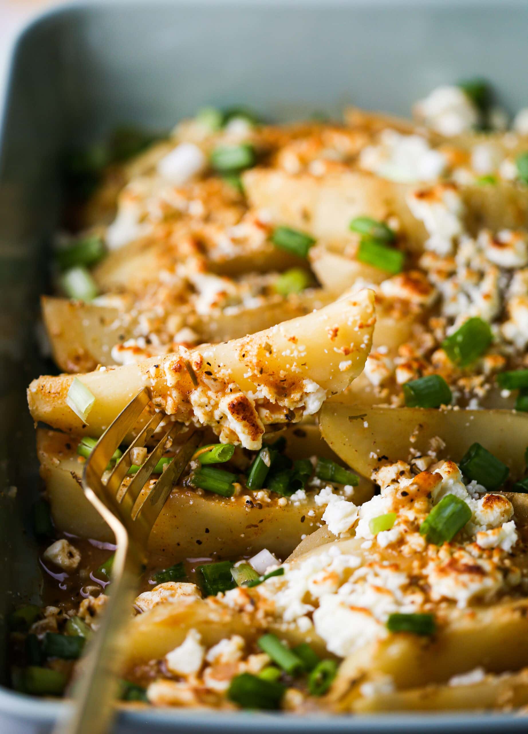 A fork holding up a lemon feta potato wedge over a potato-filled casserole dish.