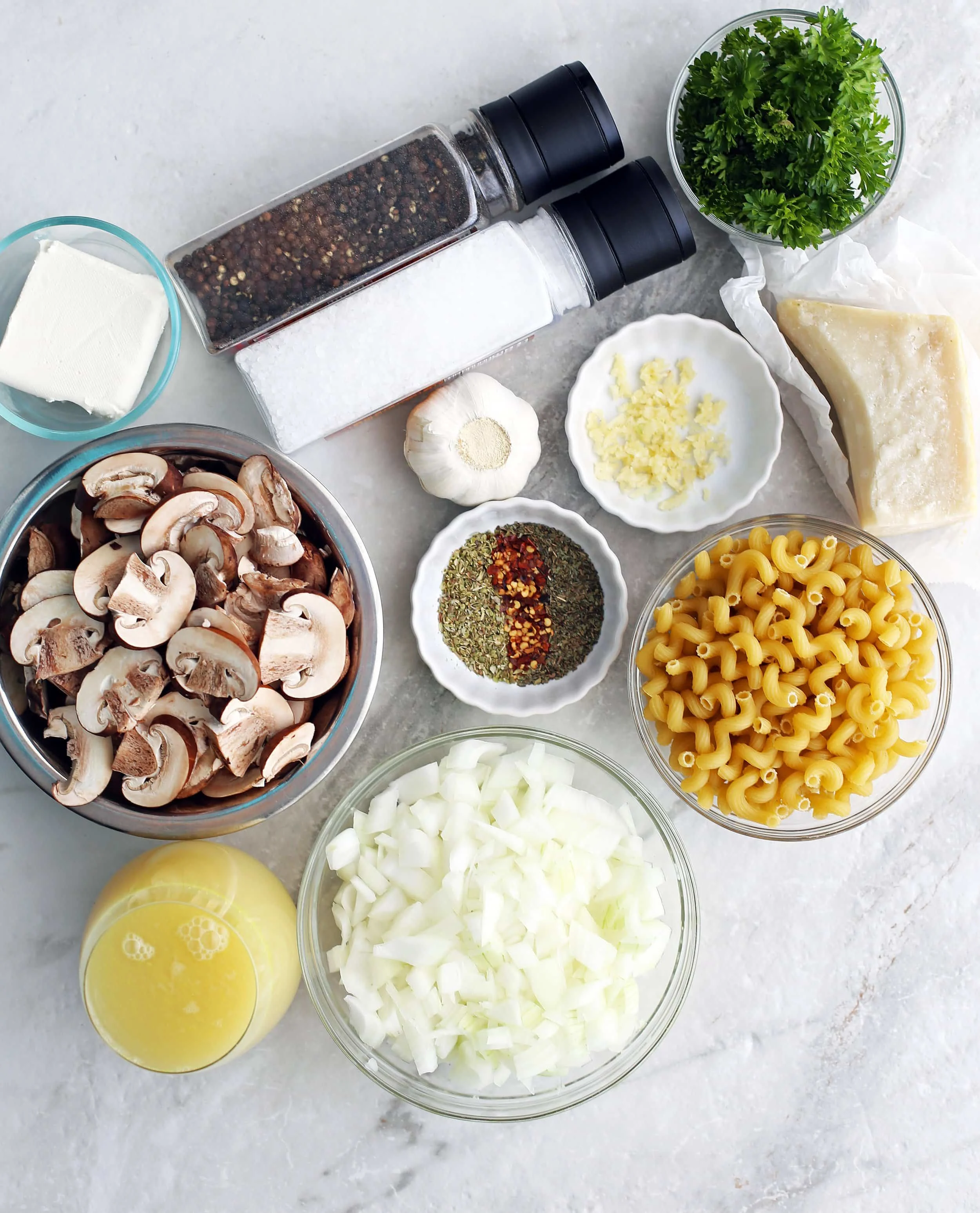 Bowls of onions, cavatappi pasta, chicken broth, mushrooms, cream cheese, parmesan and spices along with garlic, a parmesan block and salt and pepper shakers.