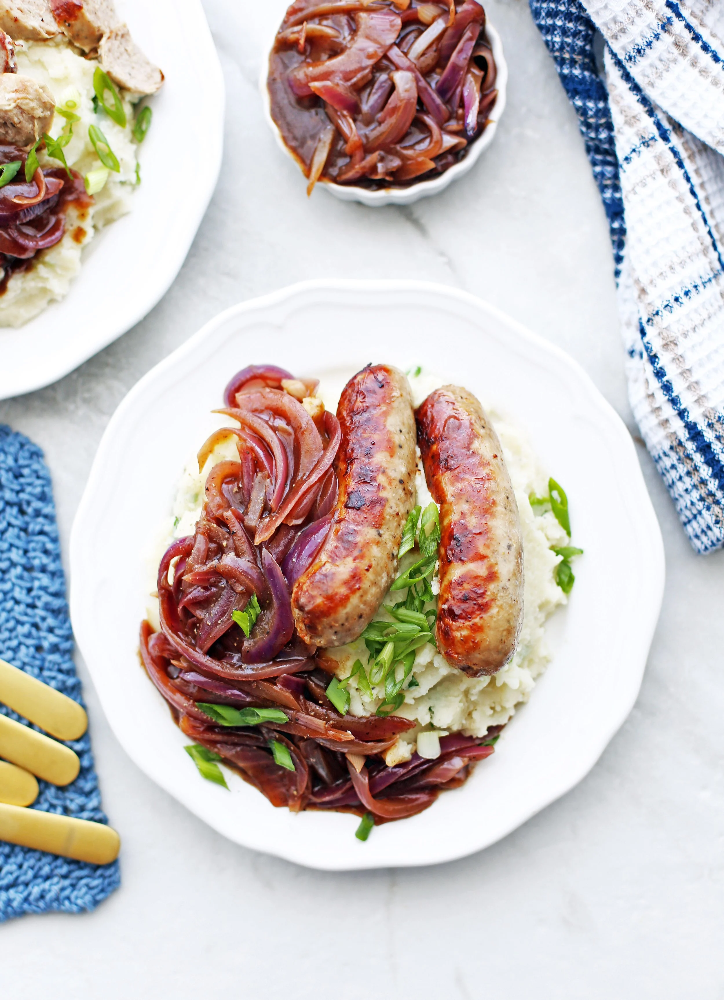 Overhead view of a heaping scoop of mashed potatoes, two pork sausages, onion gravy, and sliced green onions on a white plate.