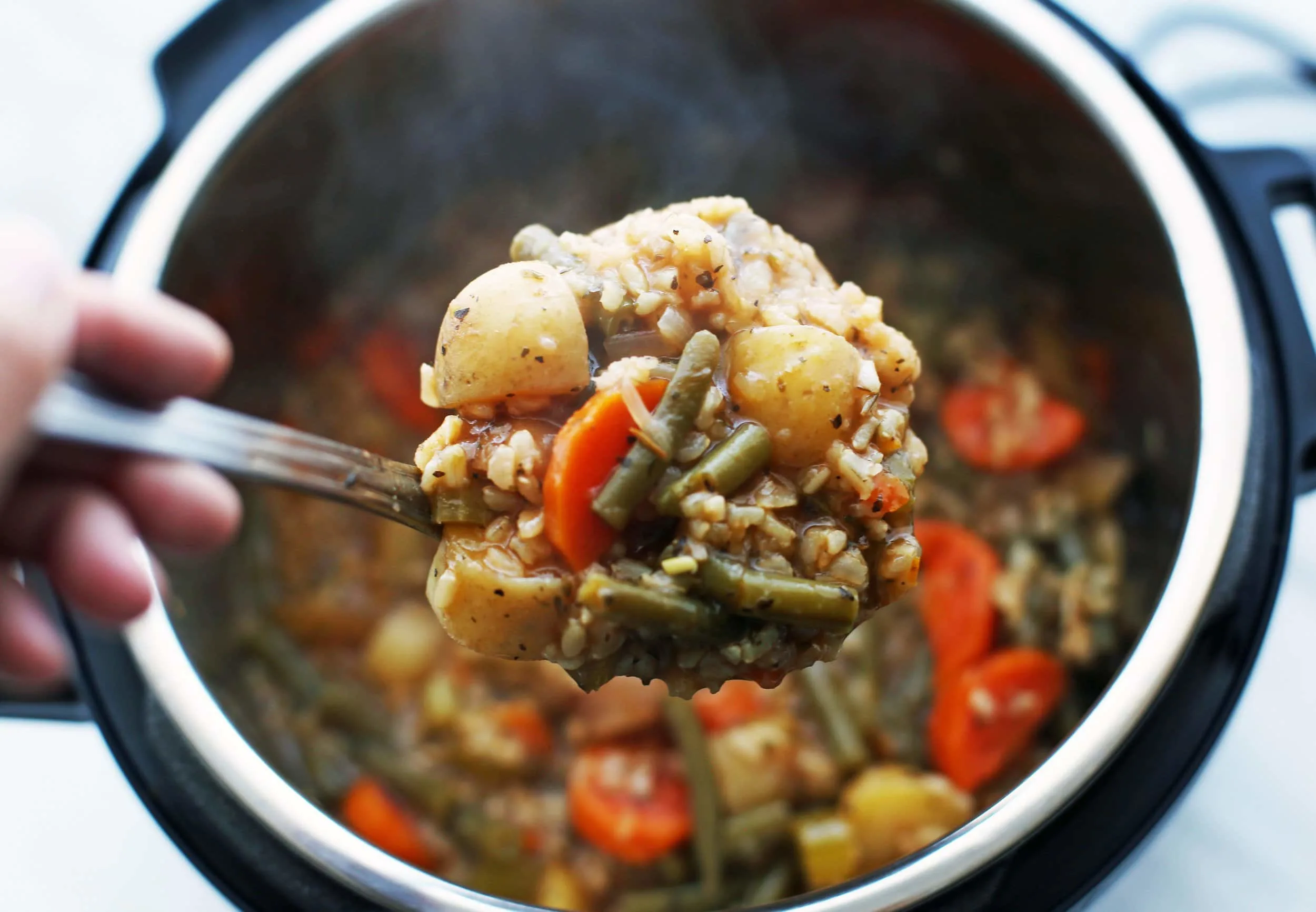 A big scoop of vegetable and brown rice soup in a metal ladle. The soup in the Instant Pot in the background.