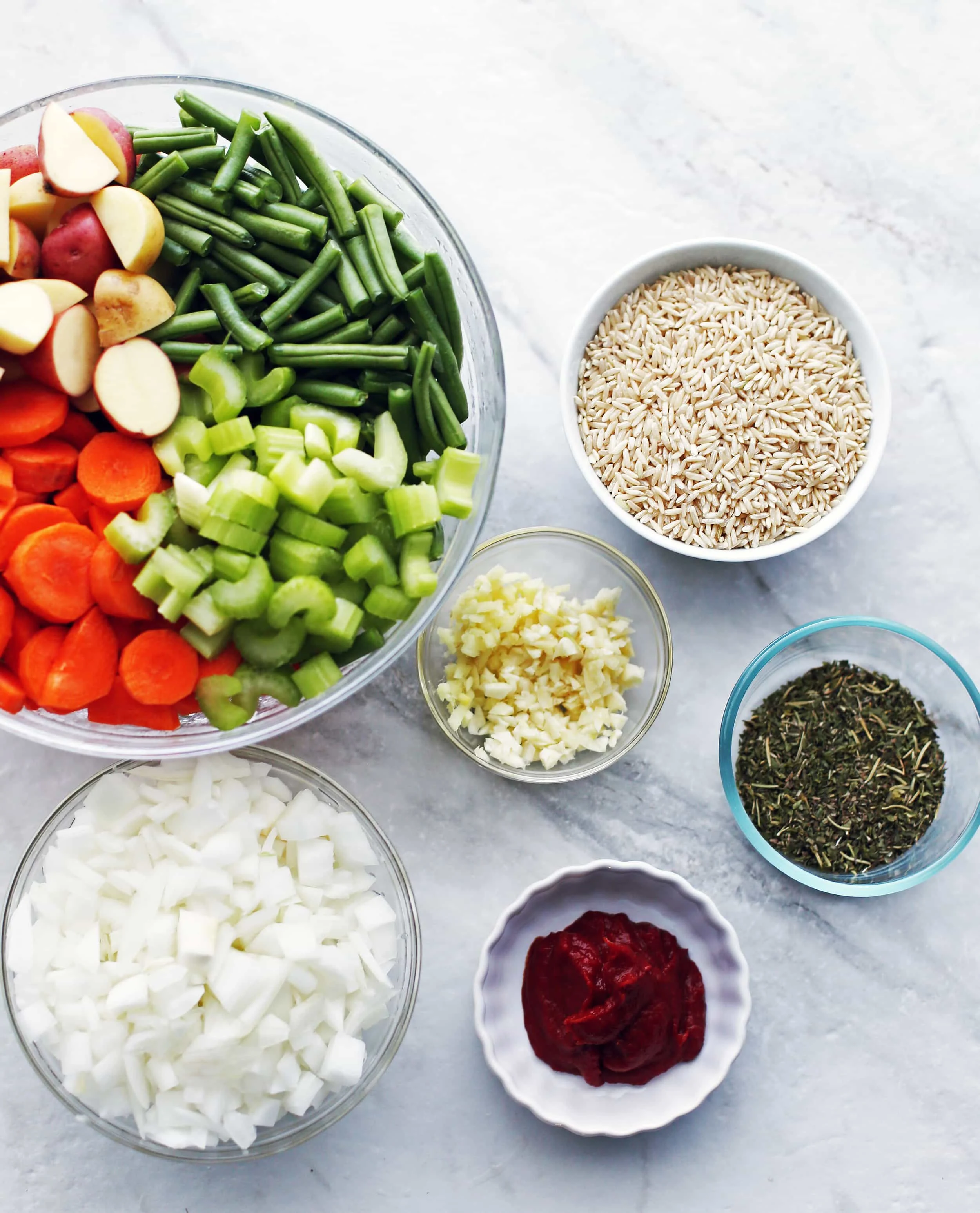 Six glass bowls with green beans, carrots, potatoes, celery, onions, garlic, ginger, brown rice, herbs, and tomato paste.