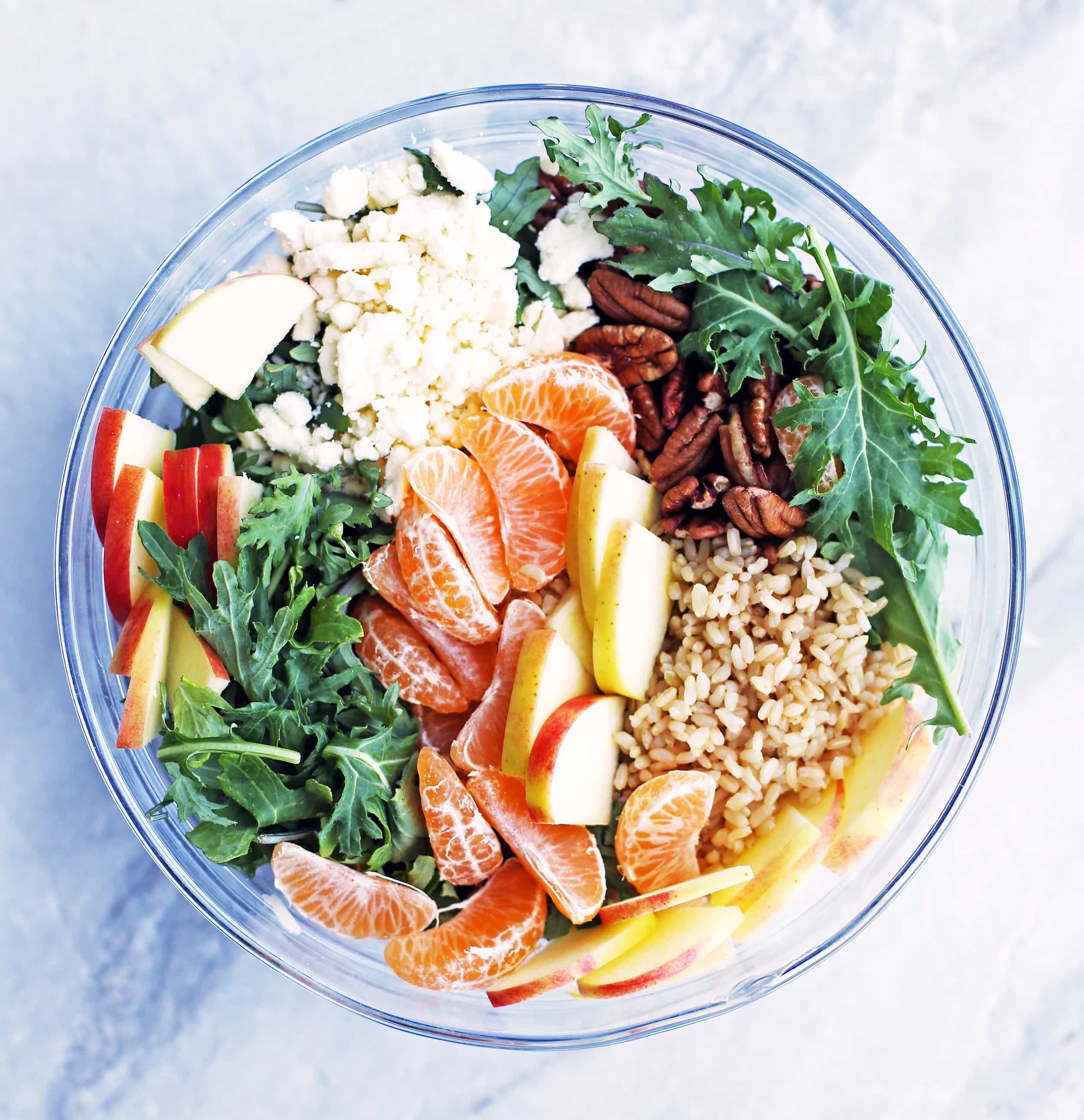 Brown rice, clementine sections, sliced apples, baby kale, and feta cheese in a large glass bowl.