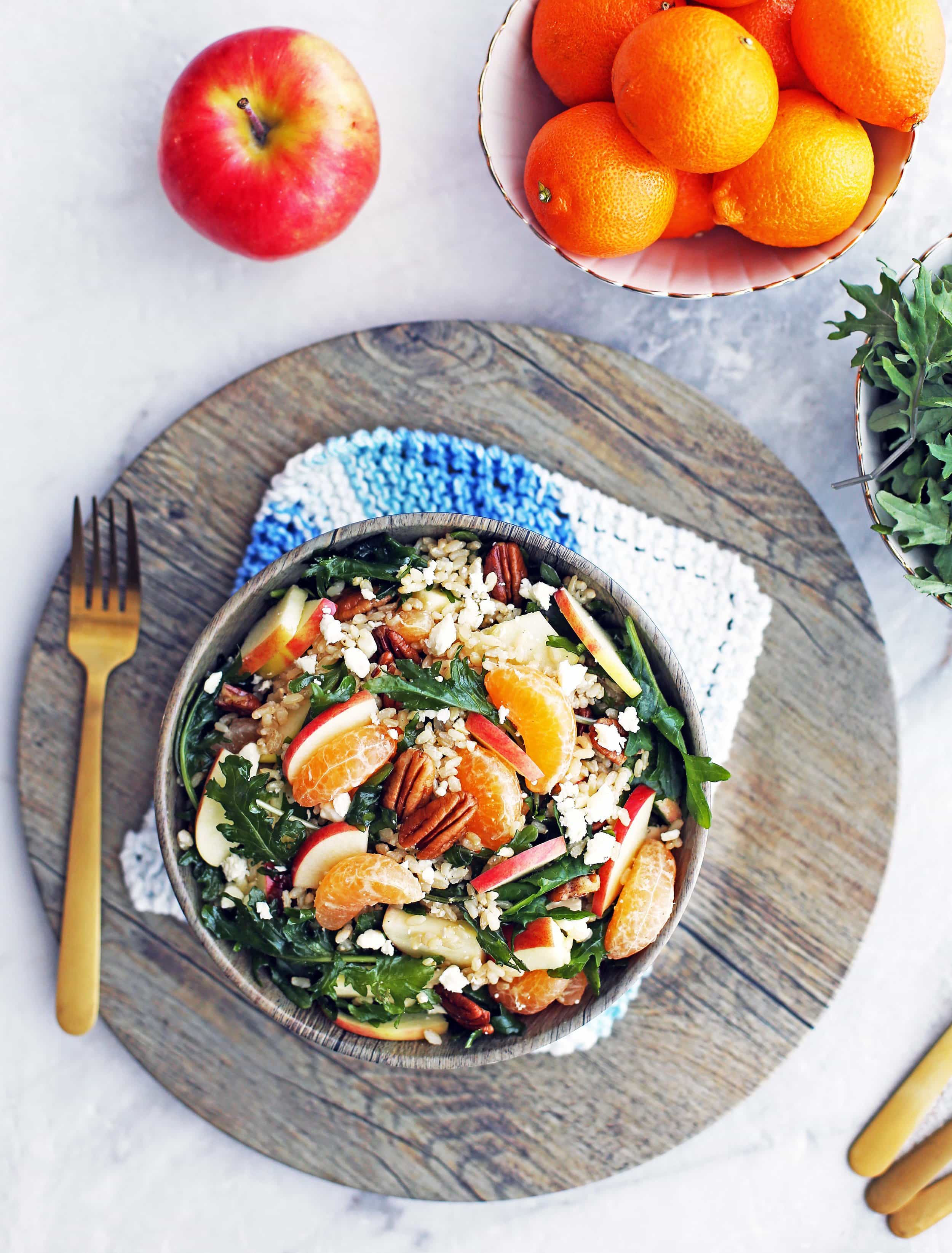 Overhead view of baby kale brown rice salad with feta, apples, and clementines in a wooden bowl, with fork to its side, on a large wooden platter.