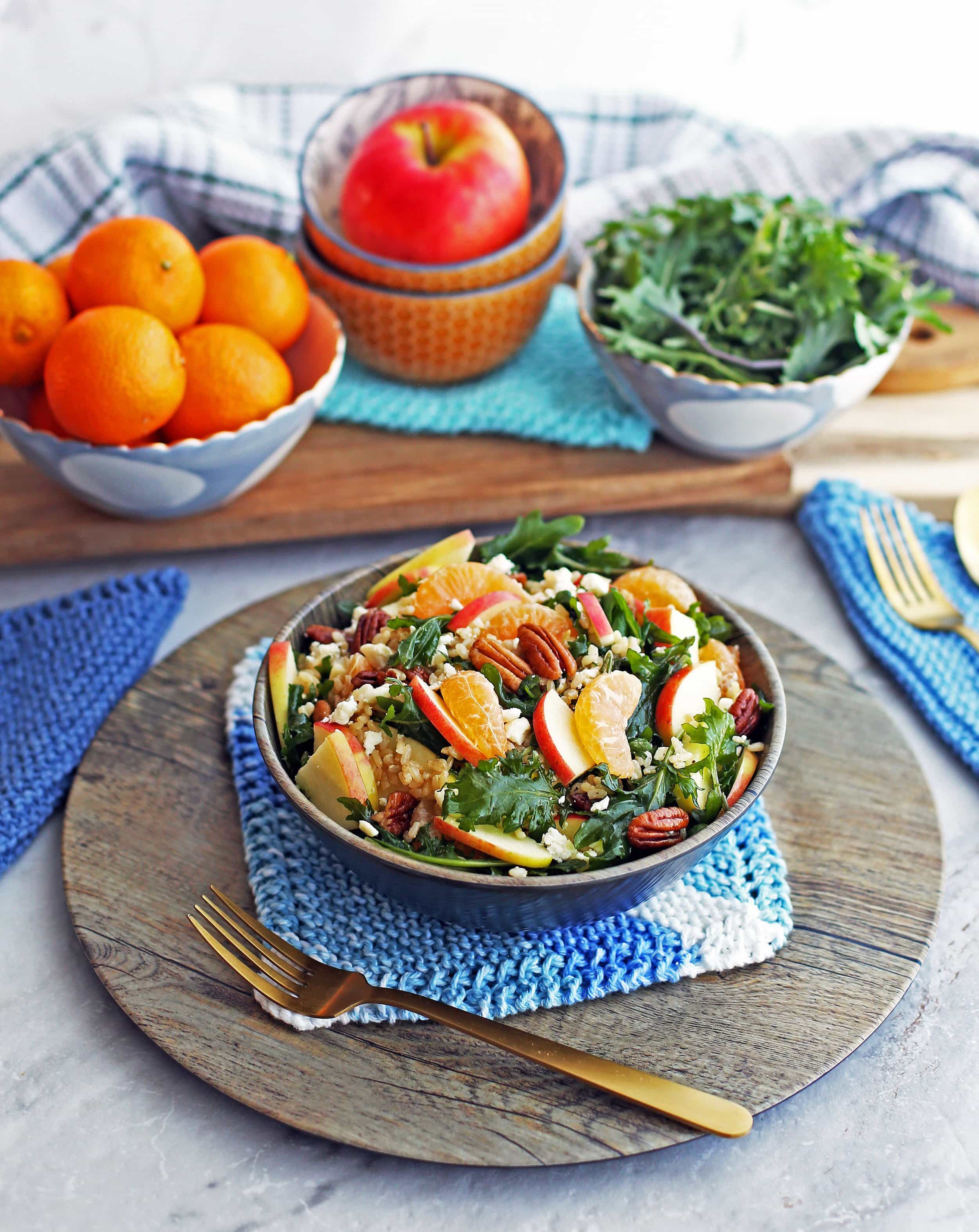 Baby kale brown rice salad with feta, apples, and clementines in a wooden bowl, a fork to the side, and bowls of clementines, apple, and baby kale in the background.