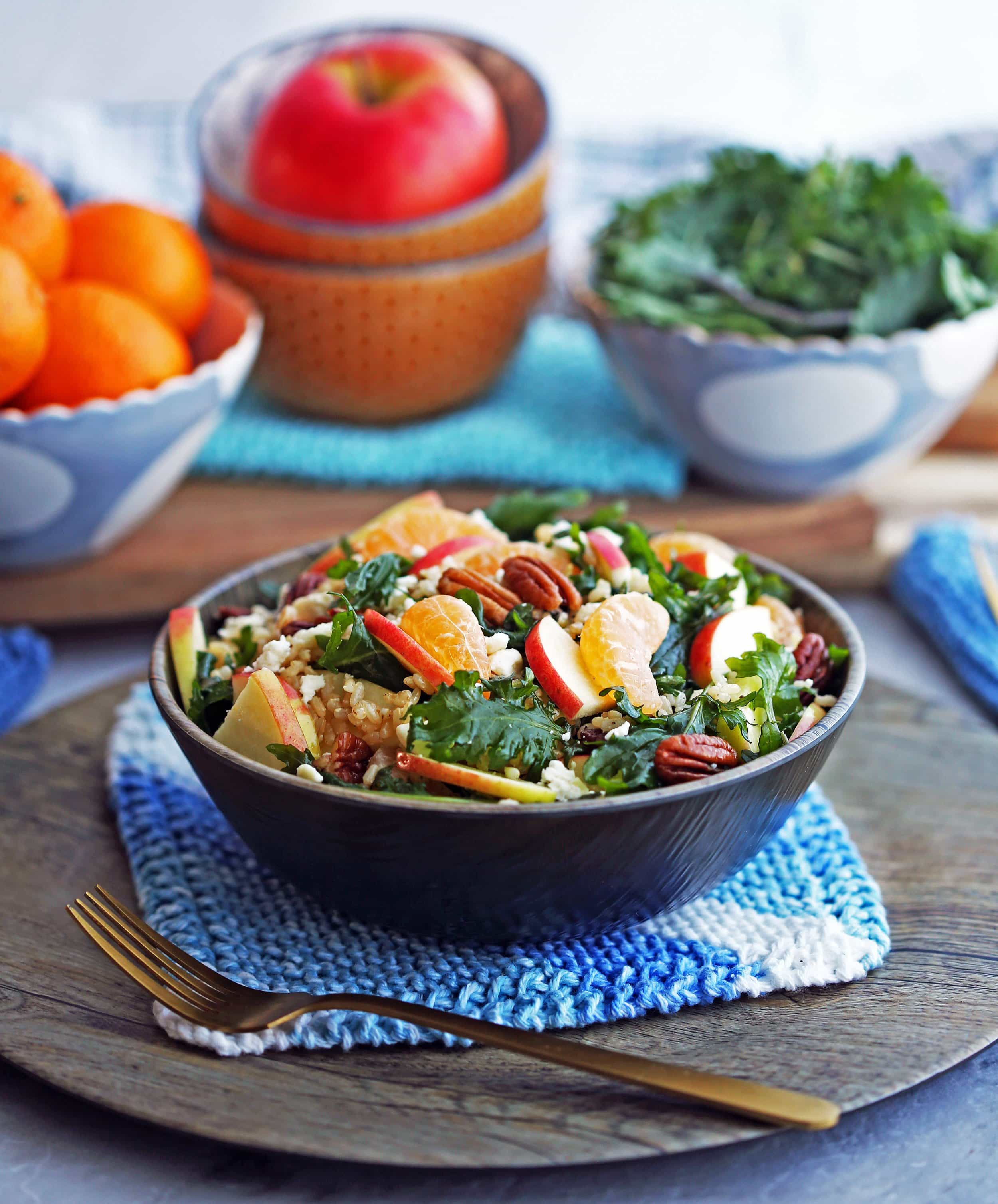 Side angled view of baby kale brown rice salad with feta, apples, and clementines in a wooden bowl, with bowls of clementines, apple, and baby kale in the background.