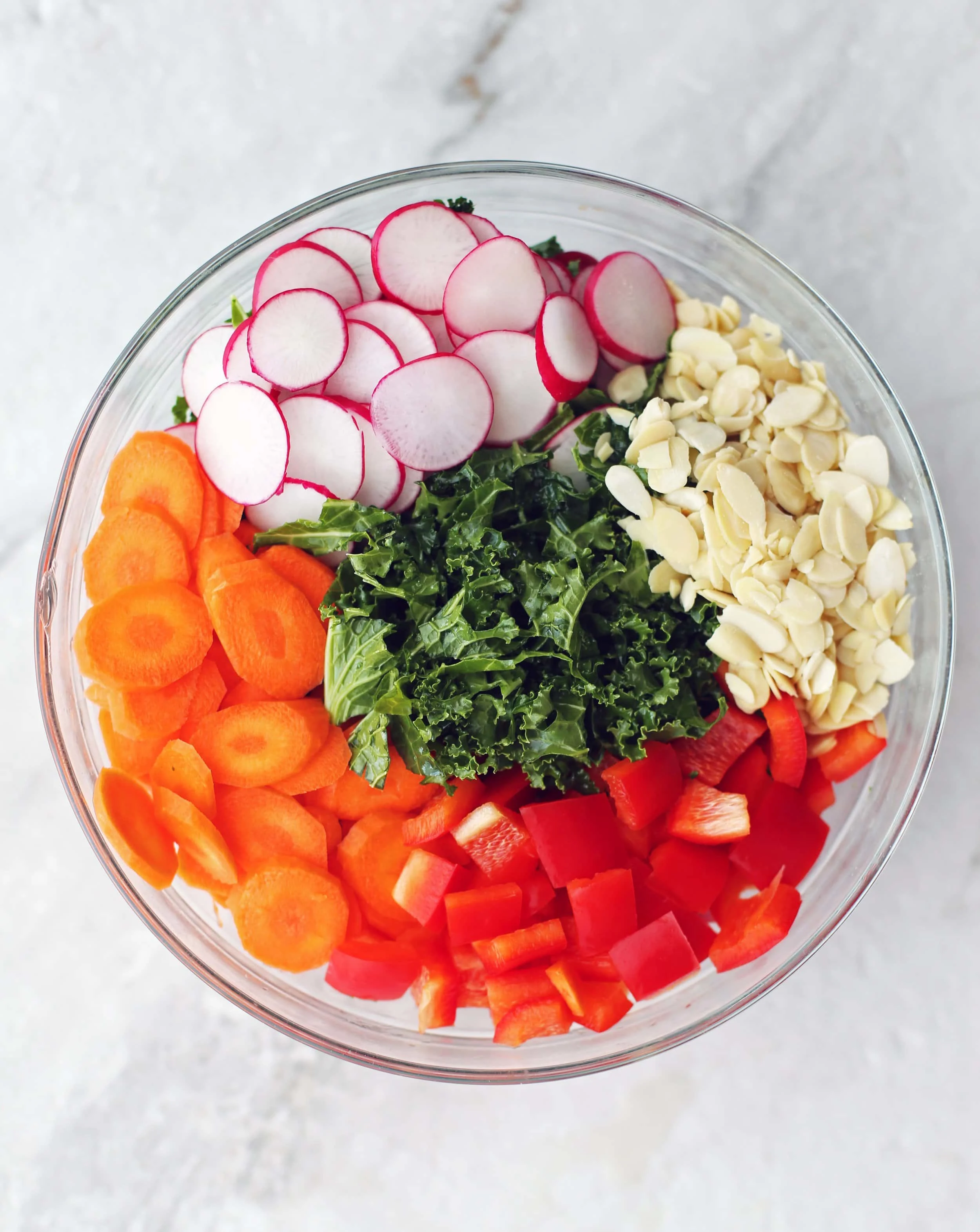 Chopped and sliced kale, carrots, bell pepper, radishes, and sliced almonds in a large glass bowl.