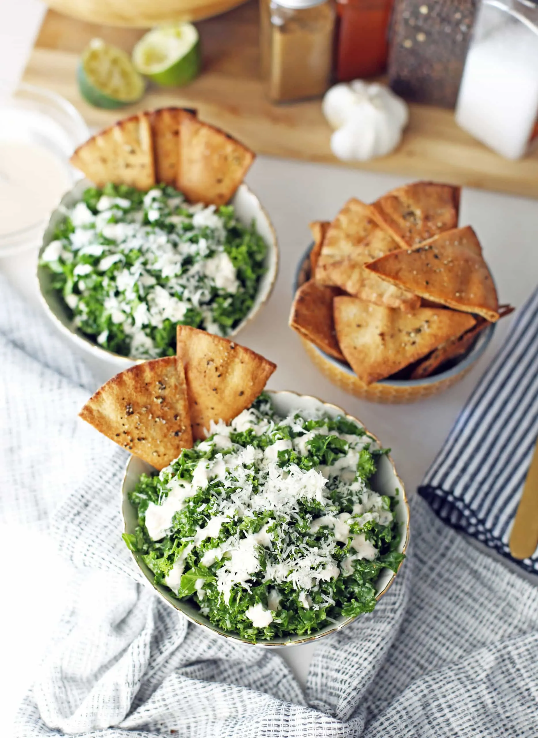 An overhead view of a small bowl of pita chips and two bowls of kale salad topped with pita chips and garlic lime dressing.