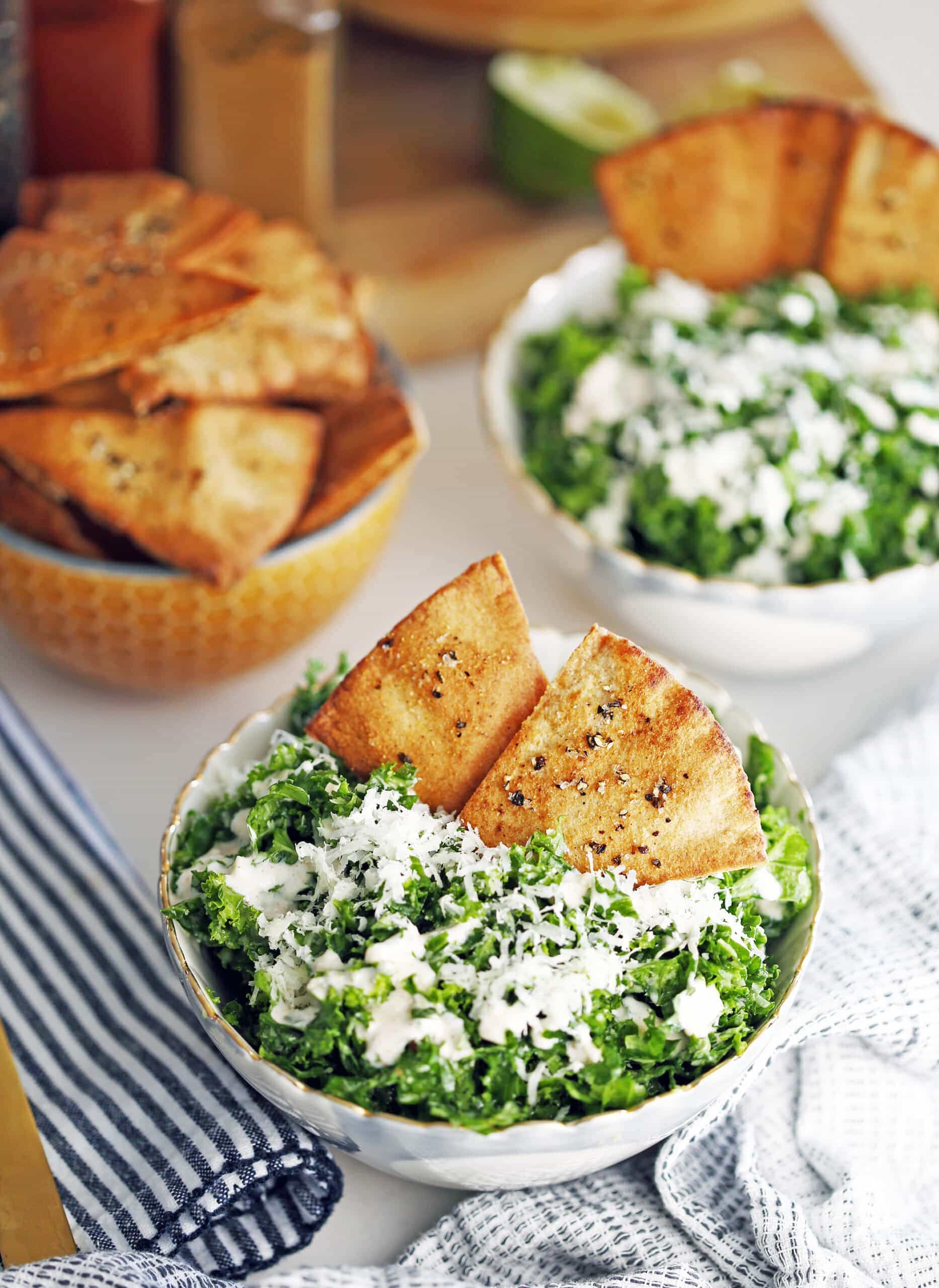 Kale parmesan salad with yogurt dressing and pita chips in two bowls and another bowl full of pita chips.