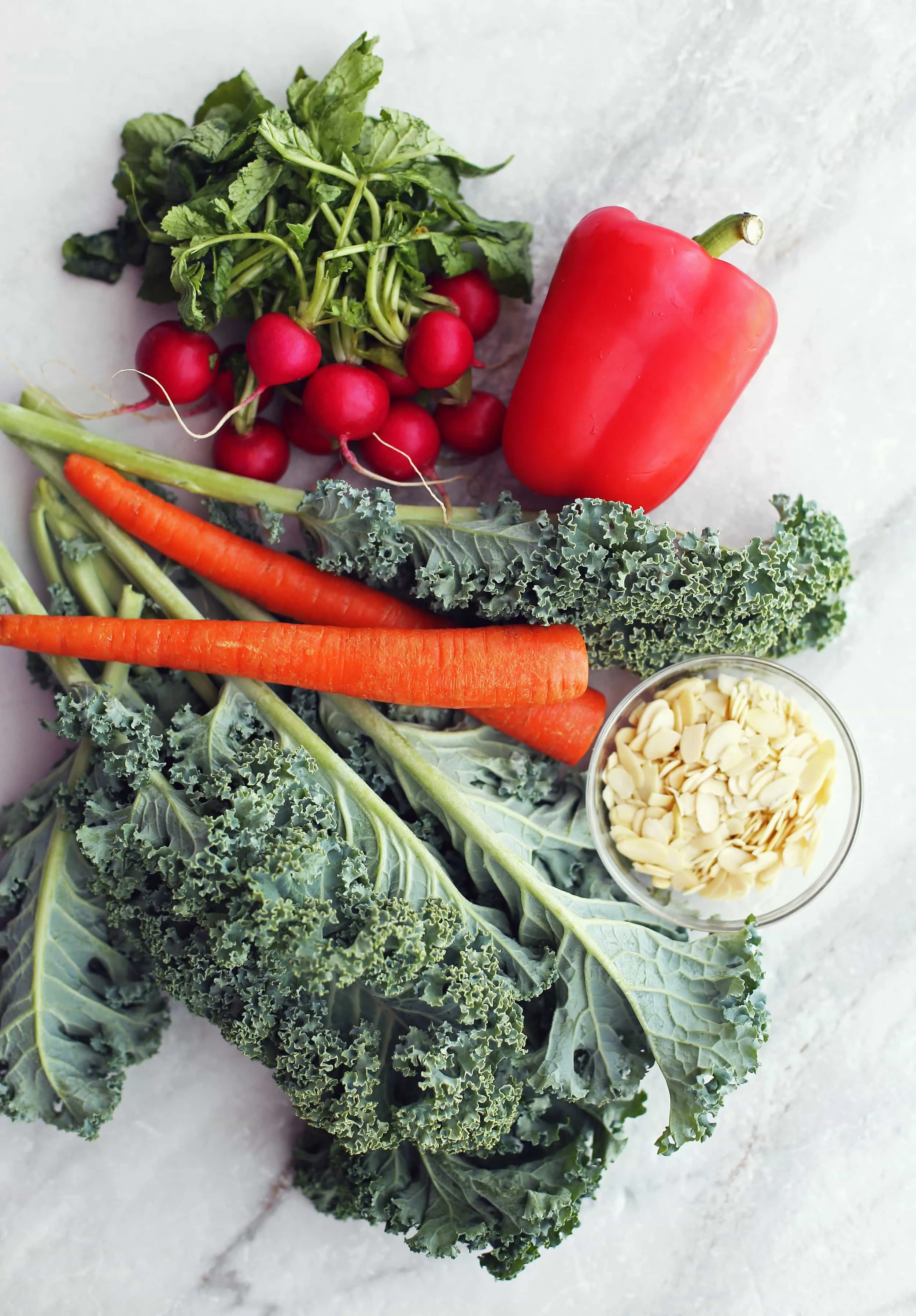 Fresh curly kale, carrots, radishes, a red bell pepper pepper and a bowl of sliced almond.