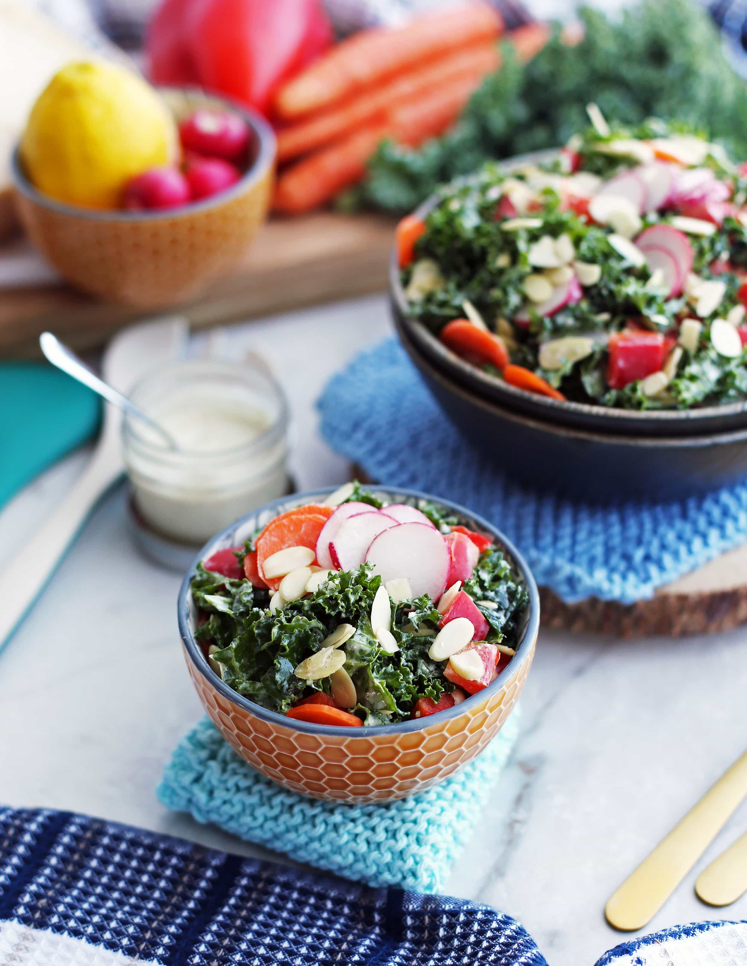 Crunchy kale, carrot, radish, and bell pepper salad with creamy parmesan yogurt dressing in a small orange bowl; more salad in a bowl behind it.