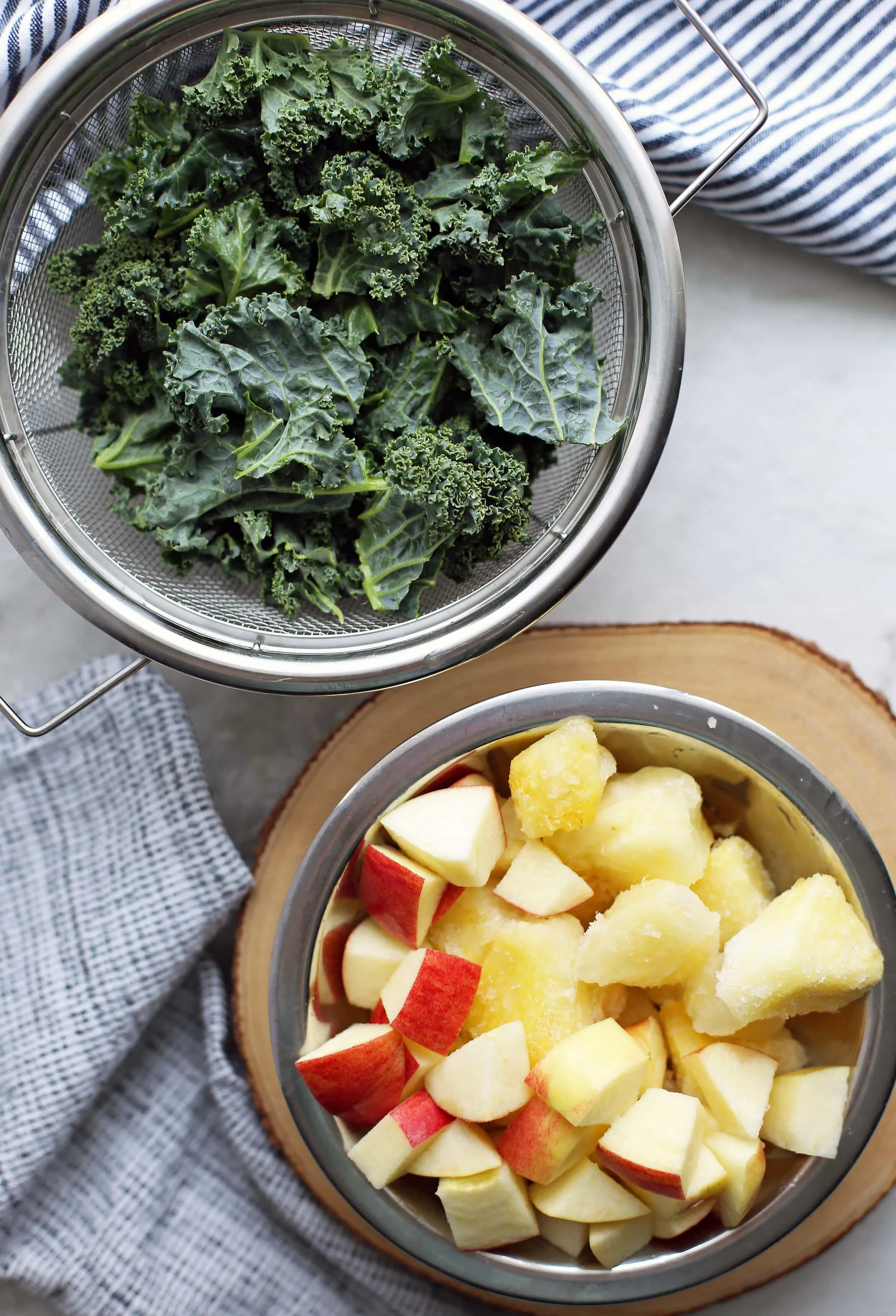 A bowl containing frozen pineapple chunks and chopped apples and a stainer containing chopped kale.