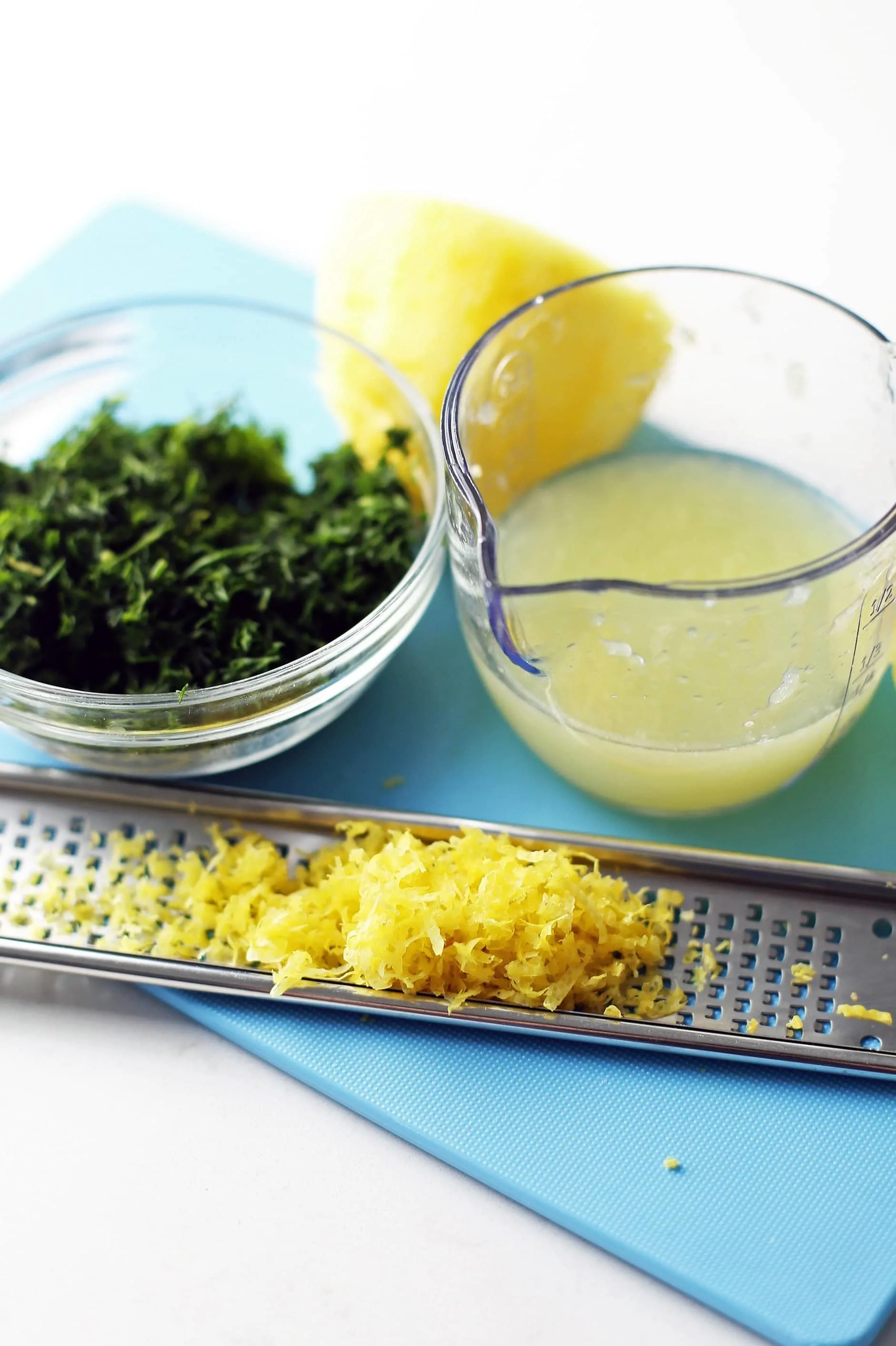 Lemon juice in a small measuring cup, lemon zest on a citrus zester, and minced fresh dill in a small glass bowl.