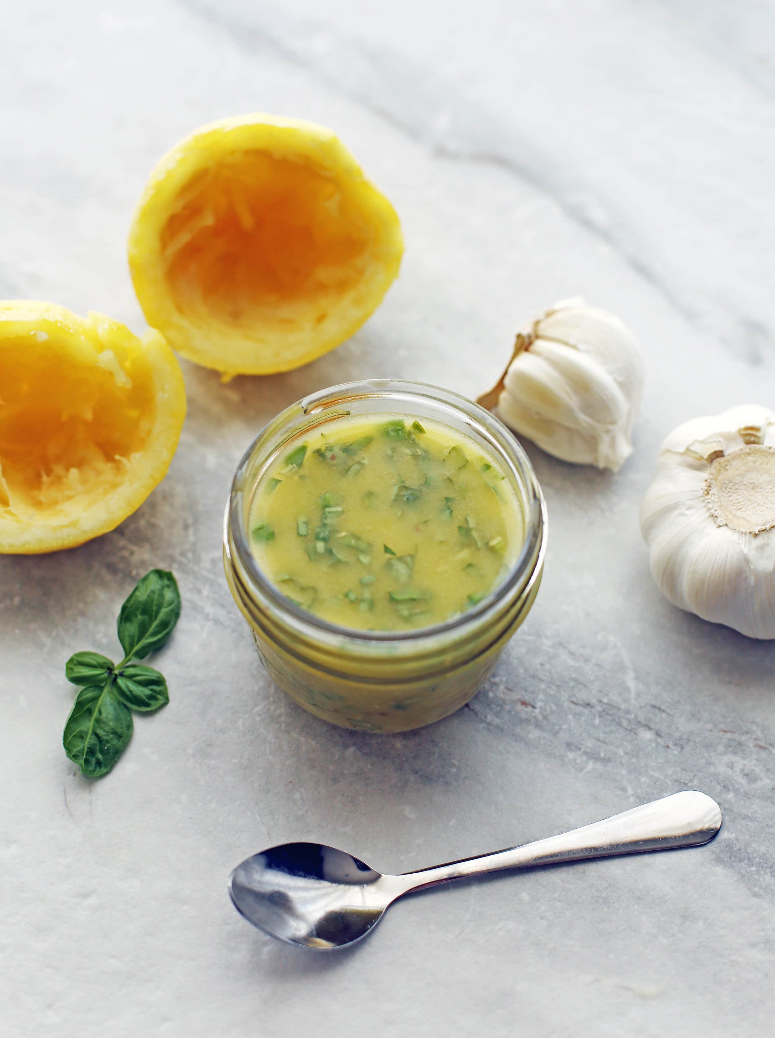 A small mason jar filled with basil lemon vinaigrette. A small spoon, basil, squeezed lemon, and garlic around it.