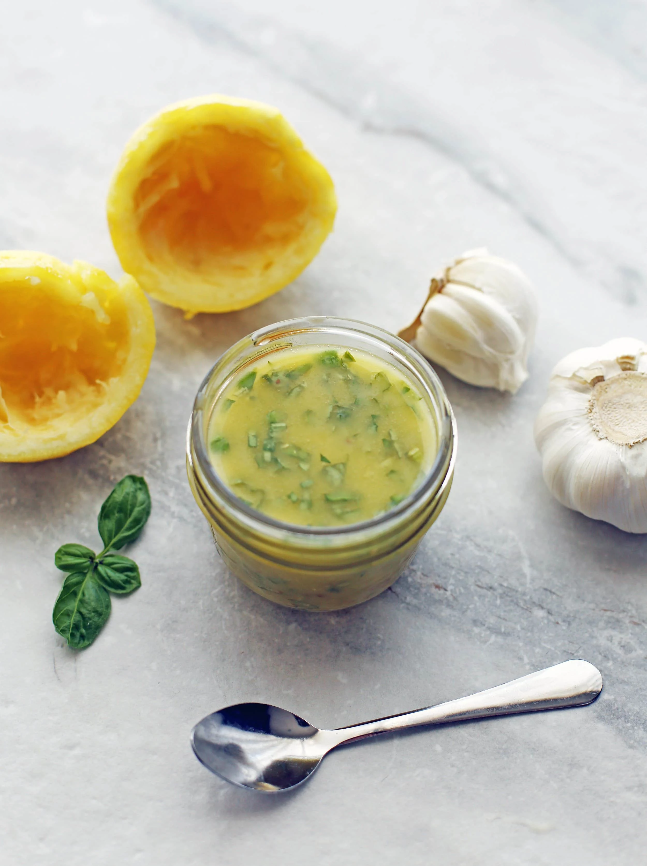 A small mason jar filled with basil lemon vinaigrette. A small spoon, basil, squeezed lemon, and garlic around it.
