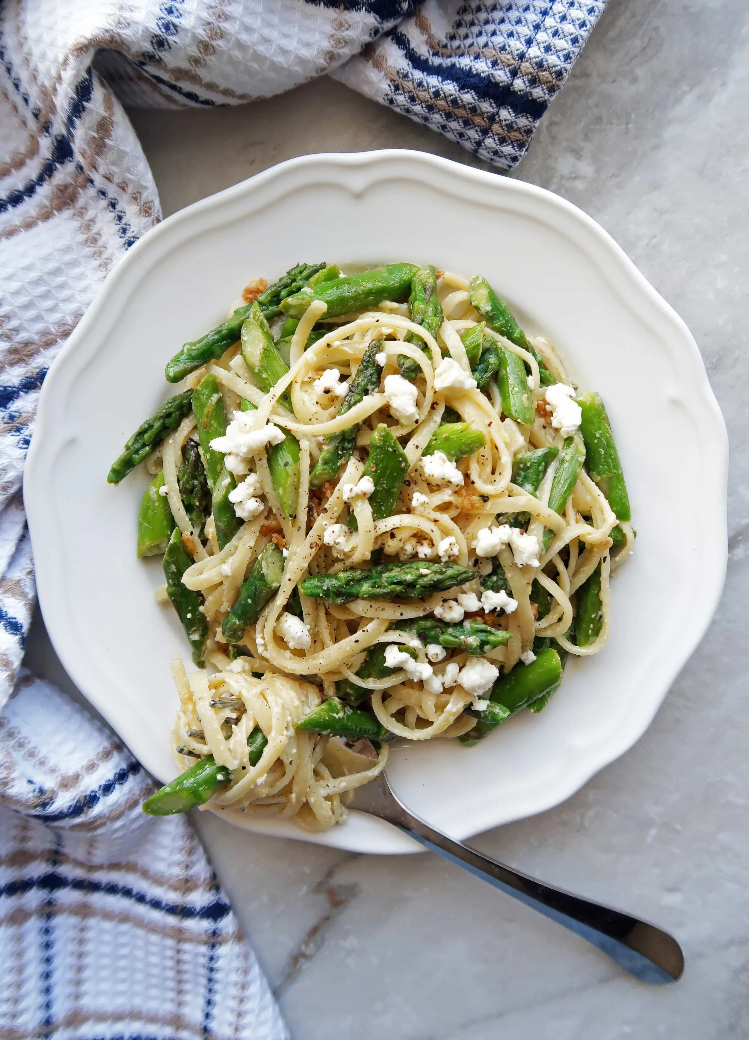 Lemon Feta Linguine with Garlic Asparagus on a white plate with linguine twirled around a fork; a kitchen towel placed beside the plate.