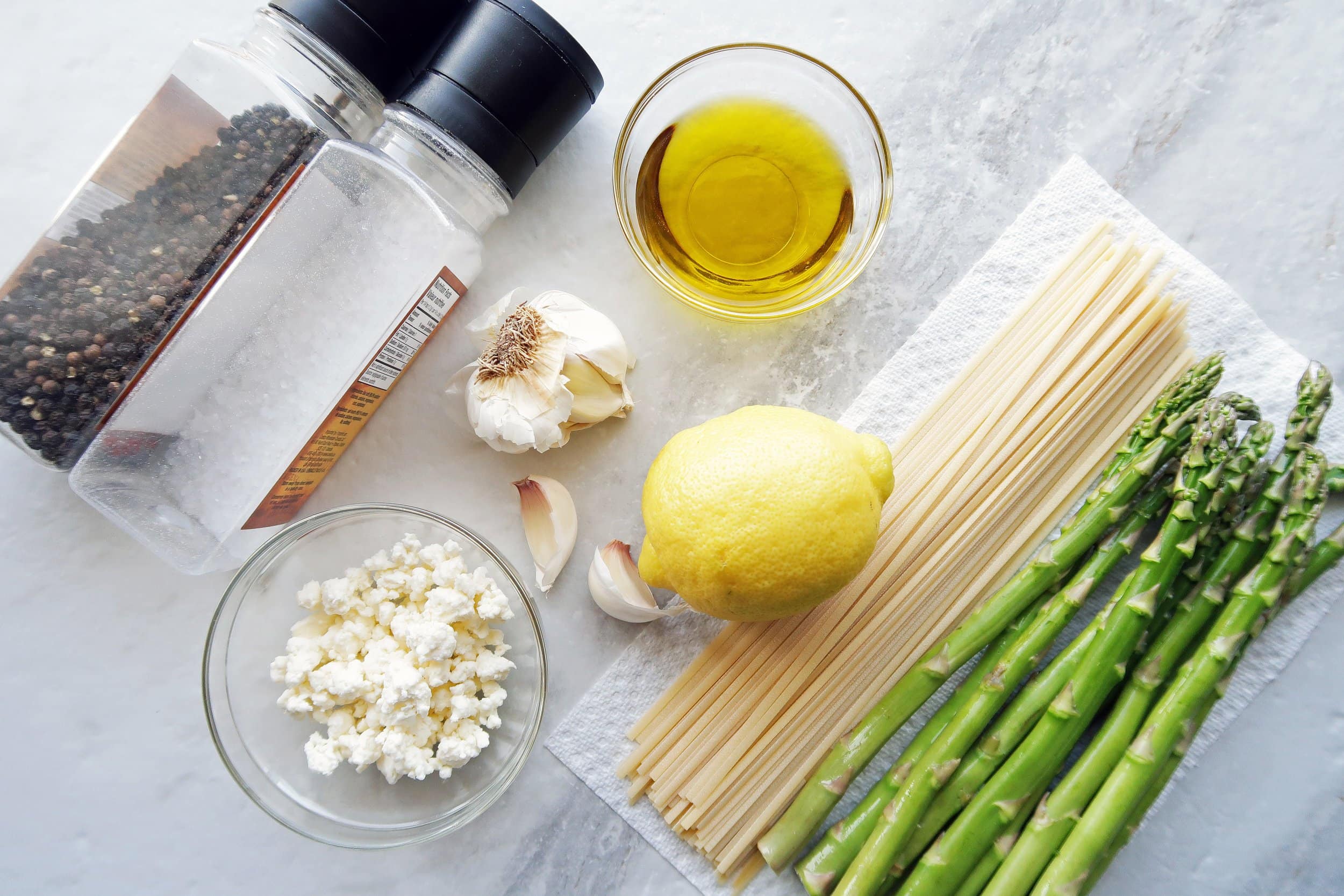 Linguine, feta, asparagus, garlic cloves, a lemon, salt and pepper shakers, and a bowl of olive oil.