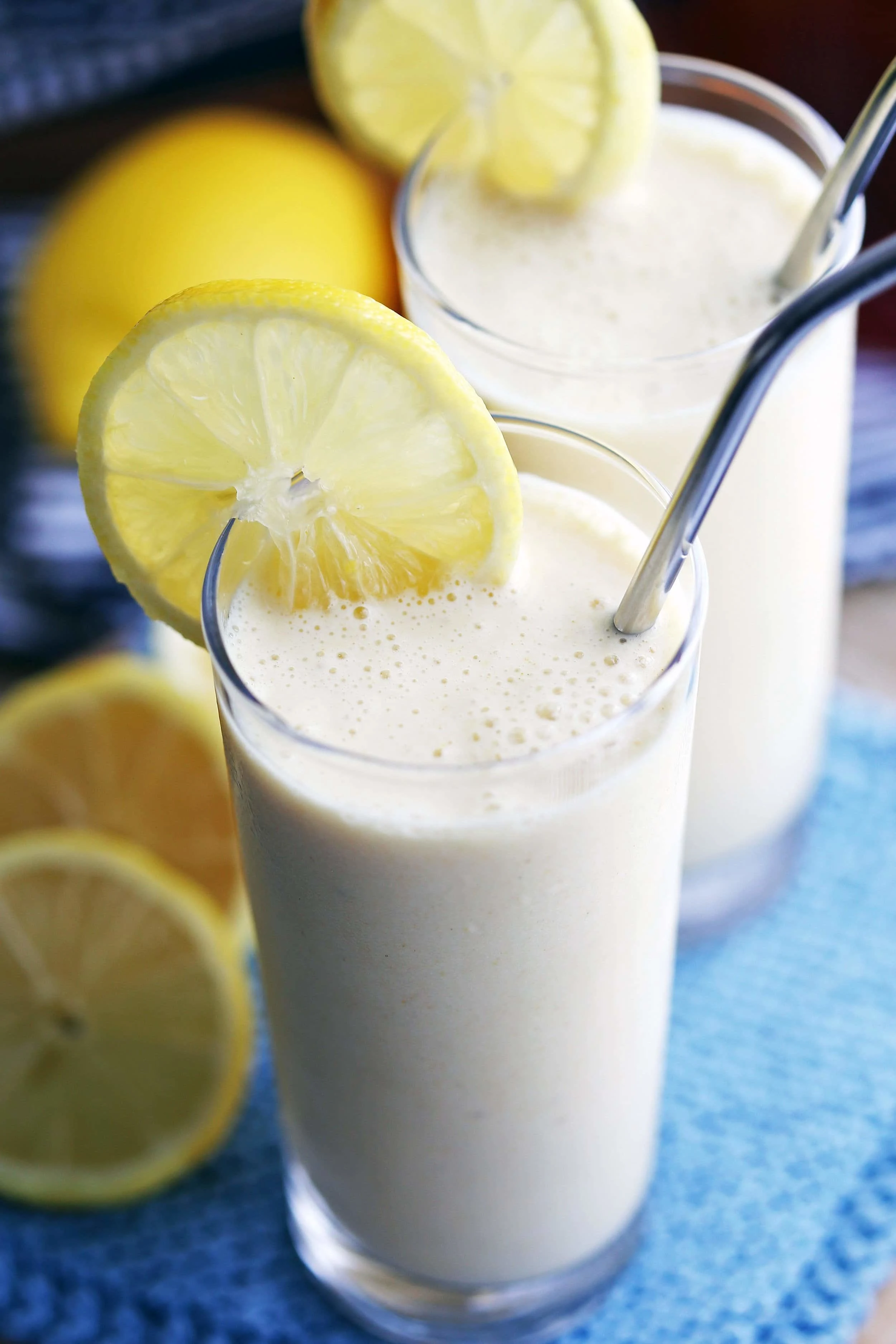 A closeup of lemon pineapple smoothie in a tall glass with metal straw and lemon garnish with another similar glass behind it.