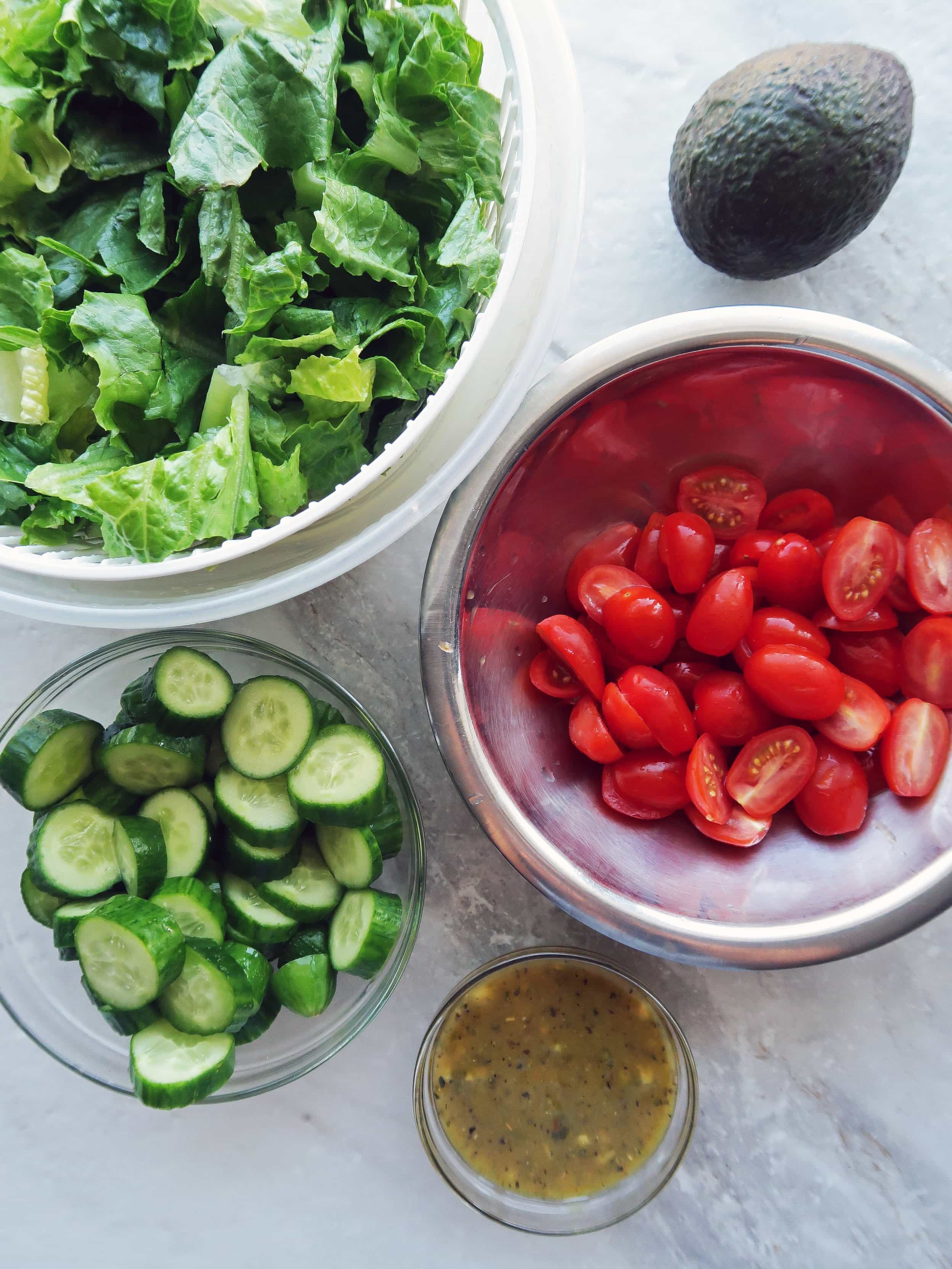 Bowls of romaine lettuce, cucumber, tomatoes, honey mustard dressing, and an avocado.