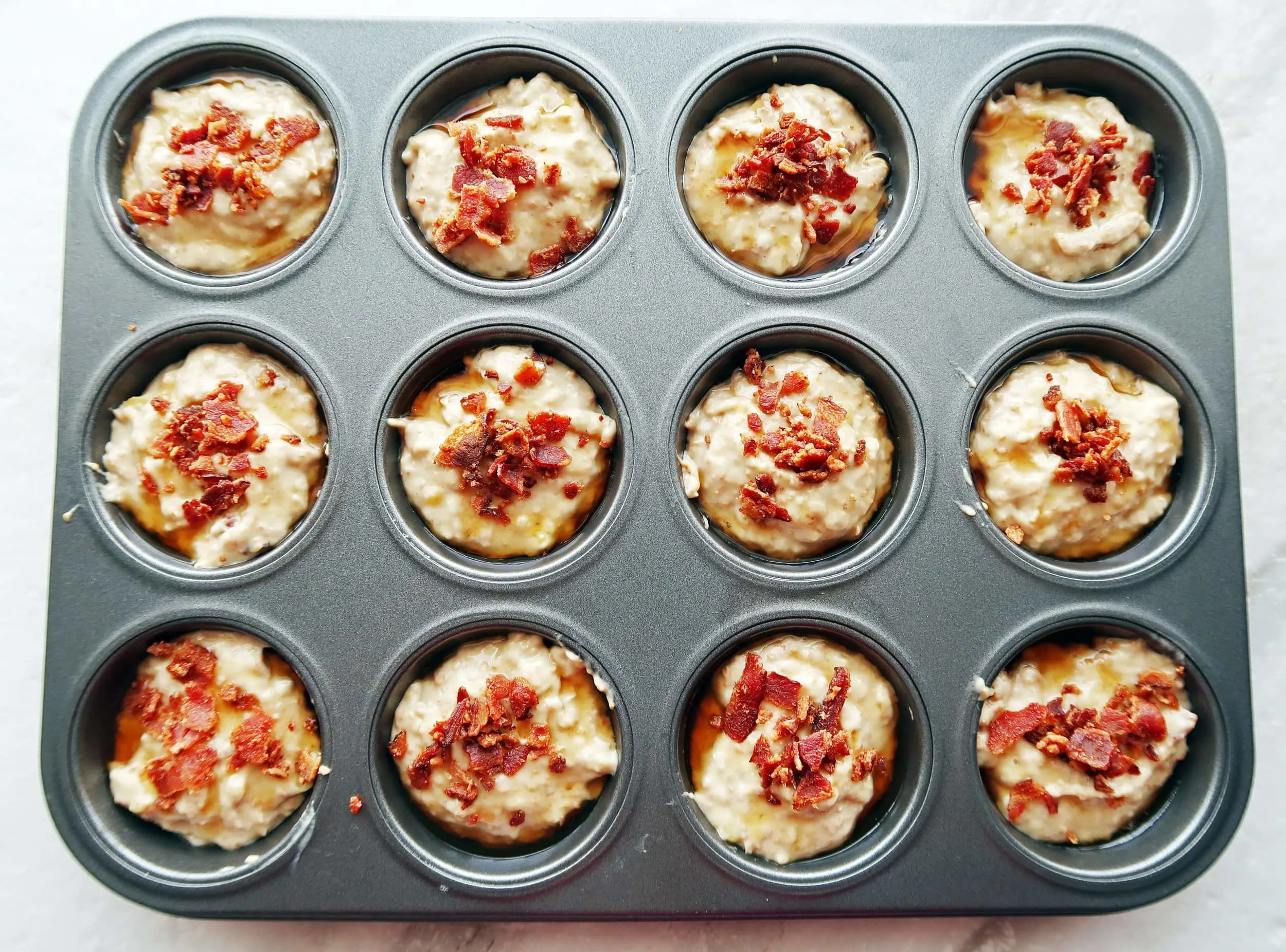 Muffin batter in a baking pan topped with bacon pieces.