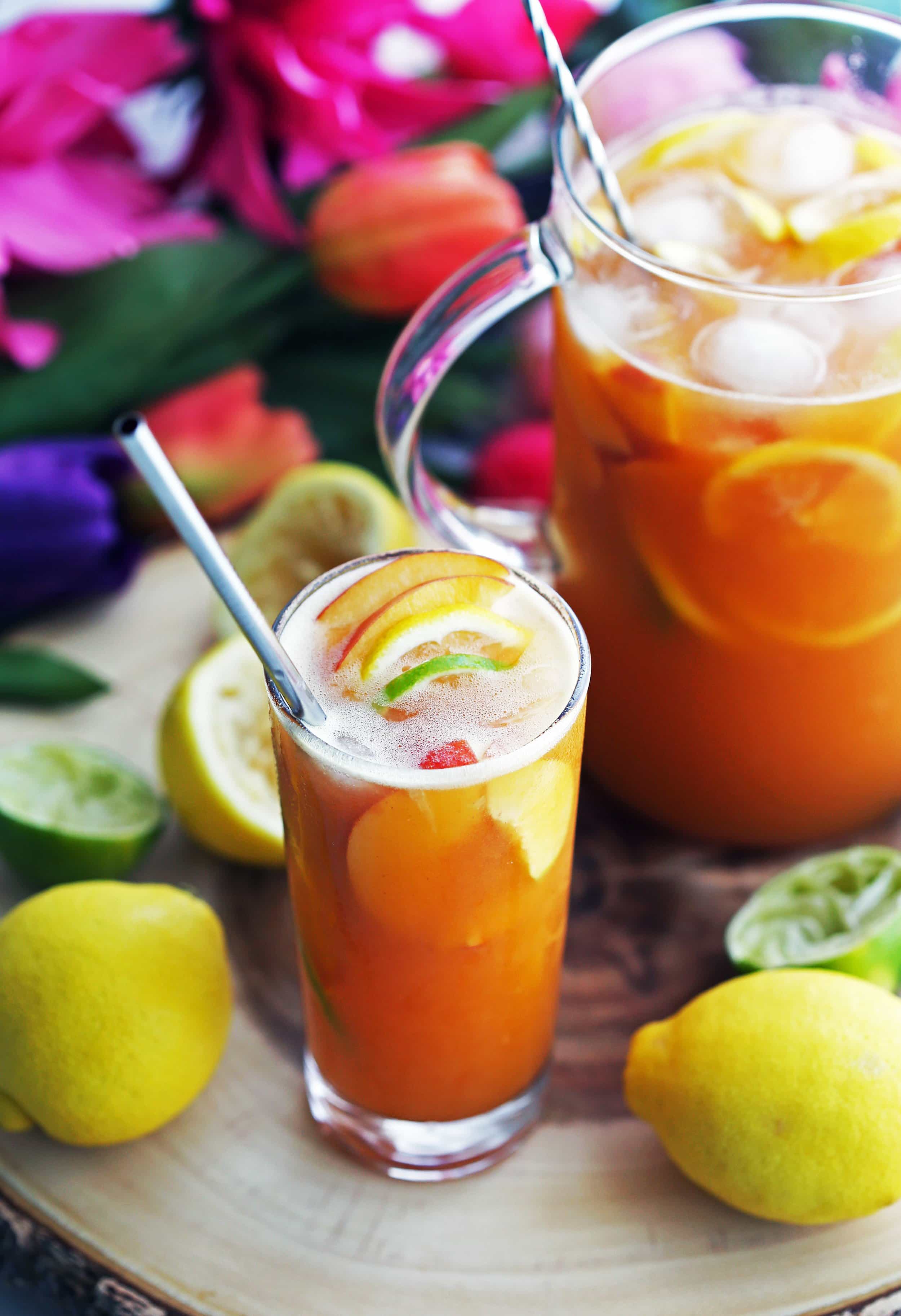 A drinking glass and glass container full of maple peach citrus juice with fruit slices and ice.