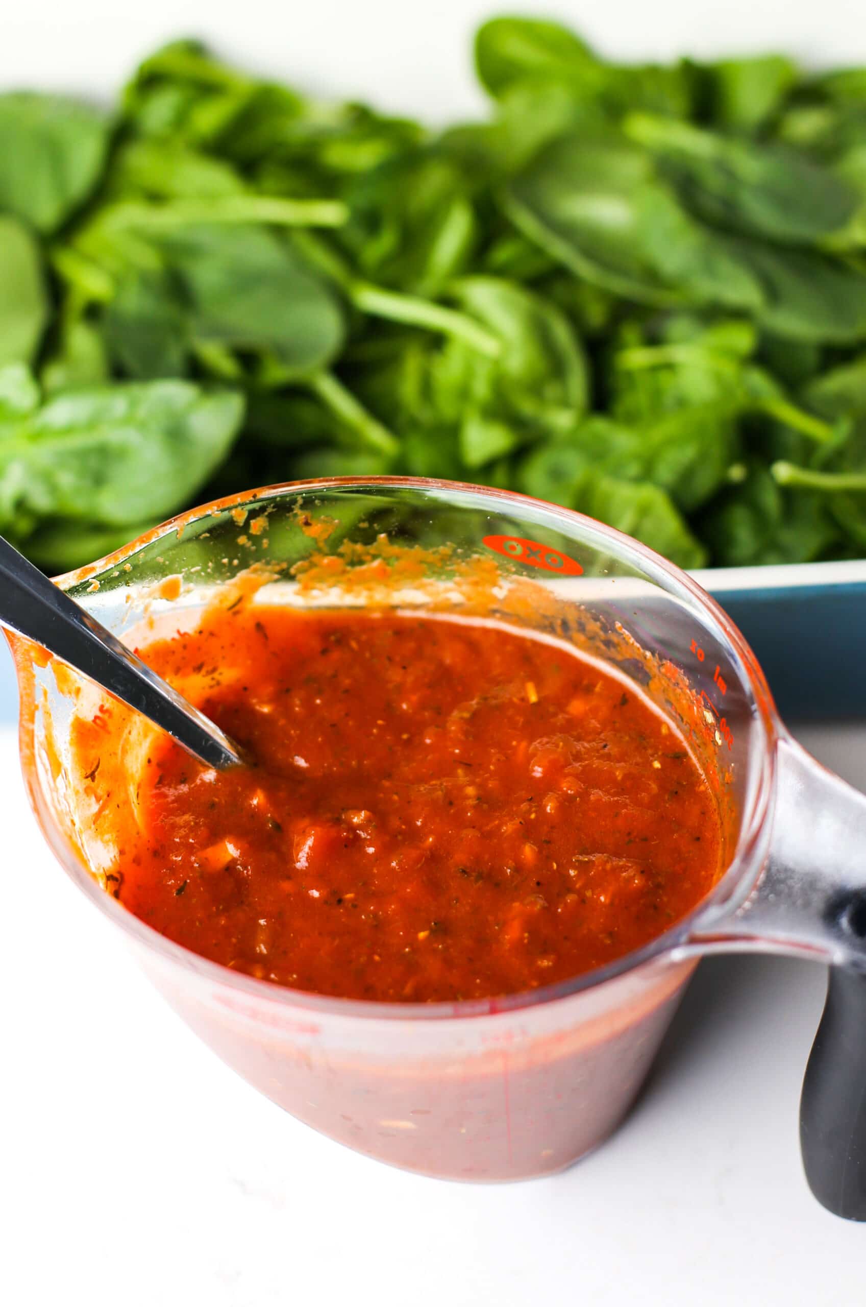 A measuring cup containing marinara sauce and a fork along with a casserole dish full of baby spinach.