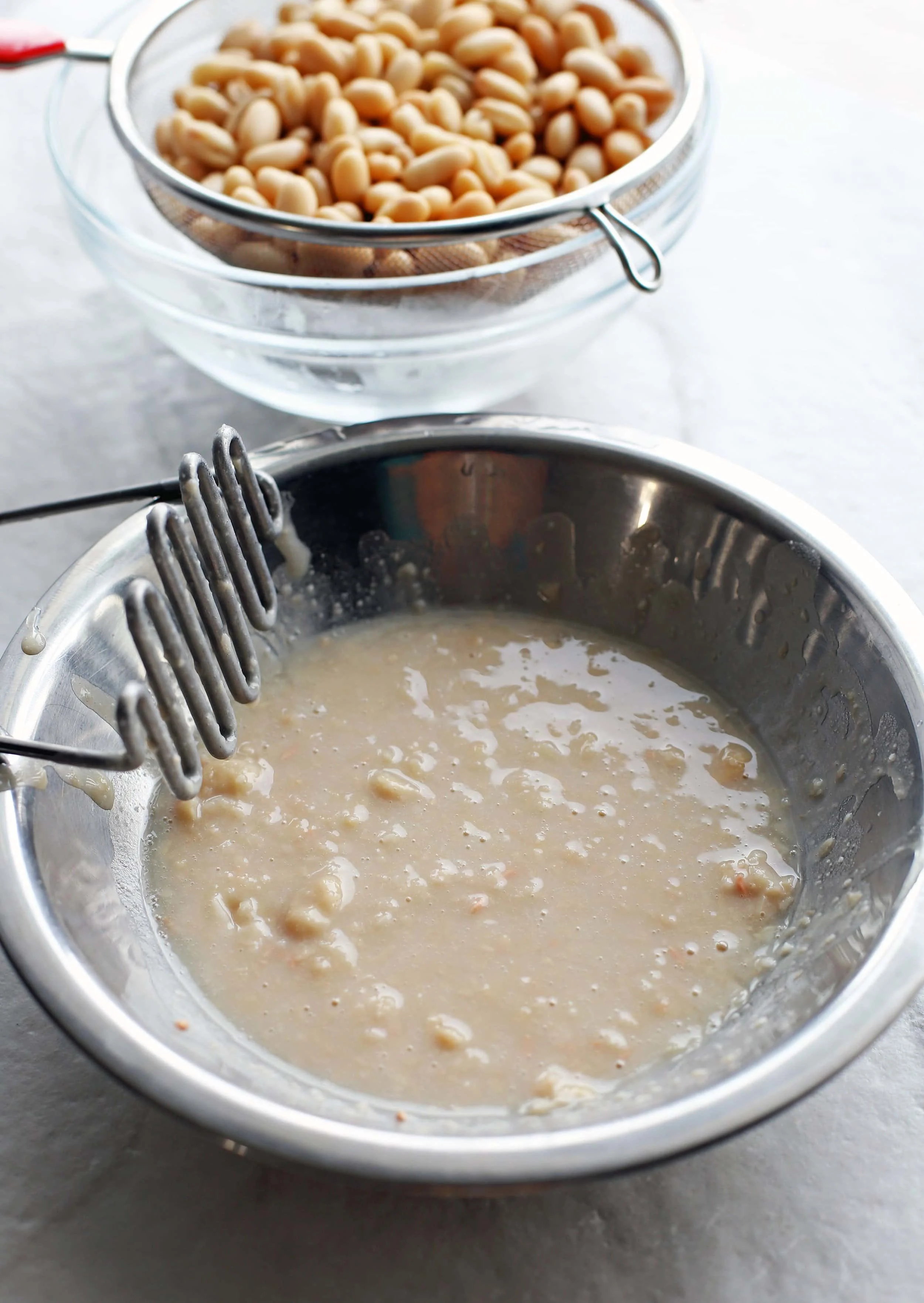 A cup of mashed white beans in one metal bowl and rinsed white beans in another bowl.