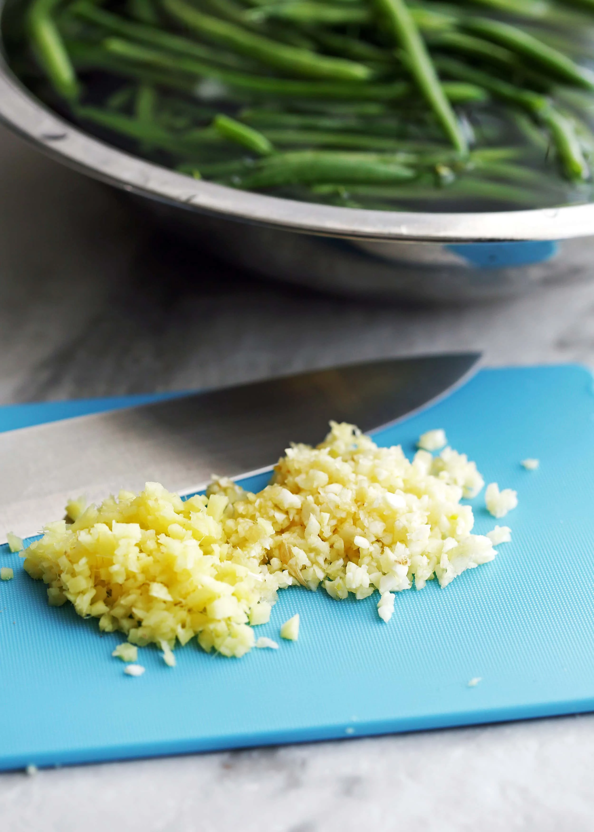 Minced garlic and ginger on a blue cutting board with a knife.