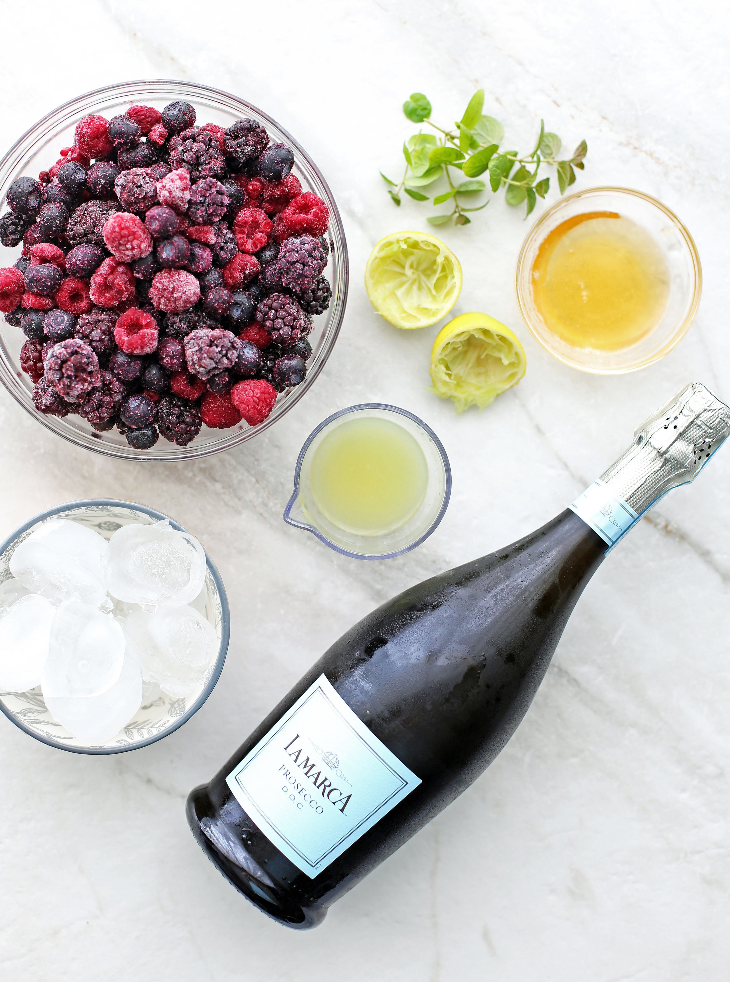 Overhead view of a bottle of Prosecco, mixed berries, ice, lime juice, fresh mint, and honey.