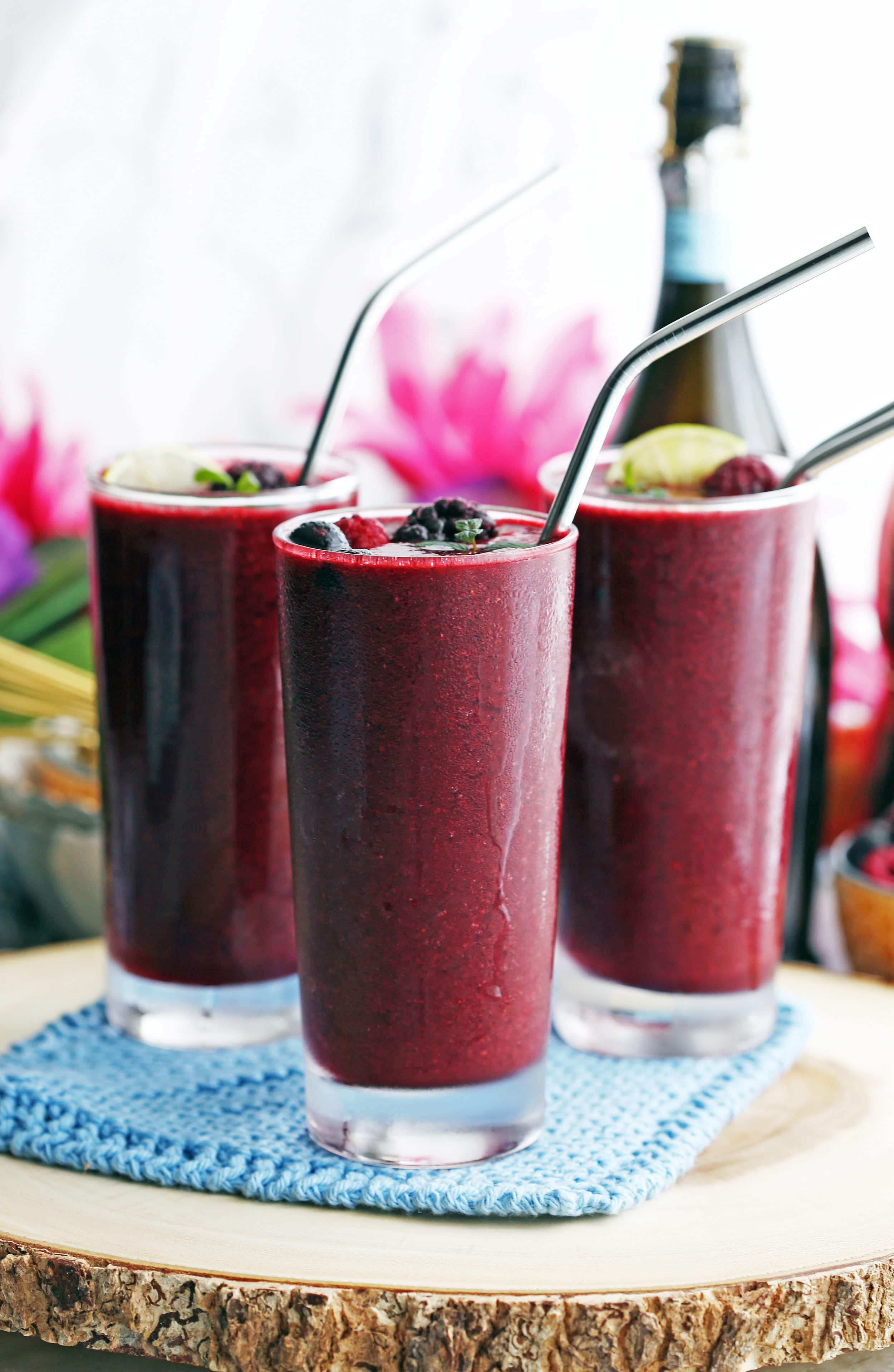 Mixed Berry Prosecco Wine Slushy drinks in three glasses with straws and topped with more berries, lime wedges, and mint.