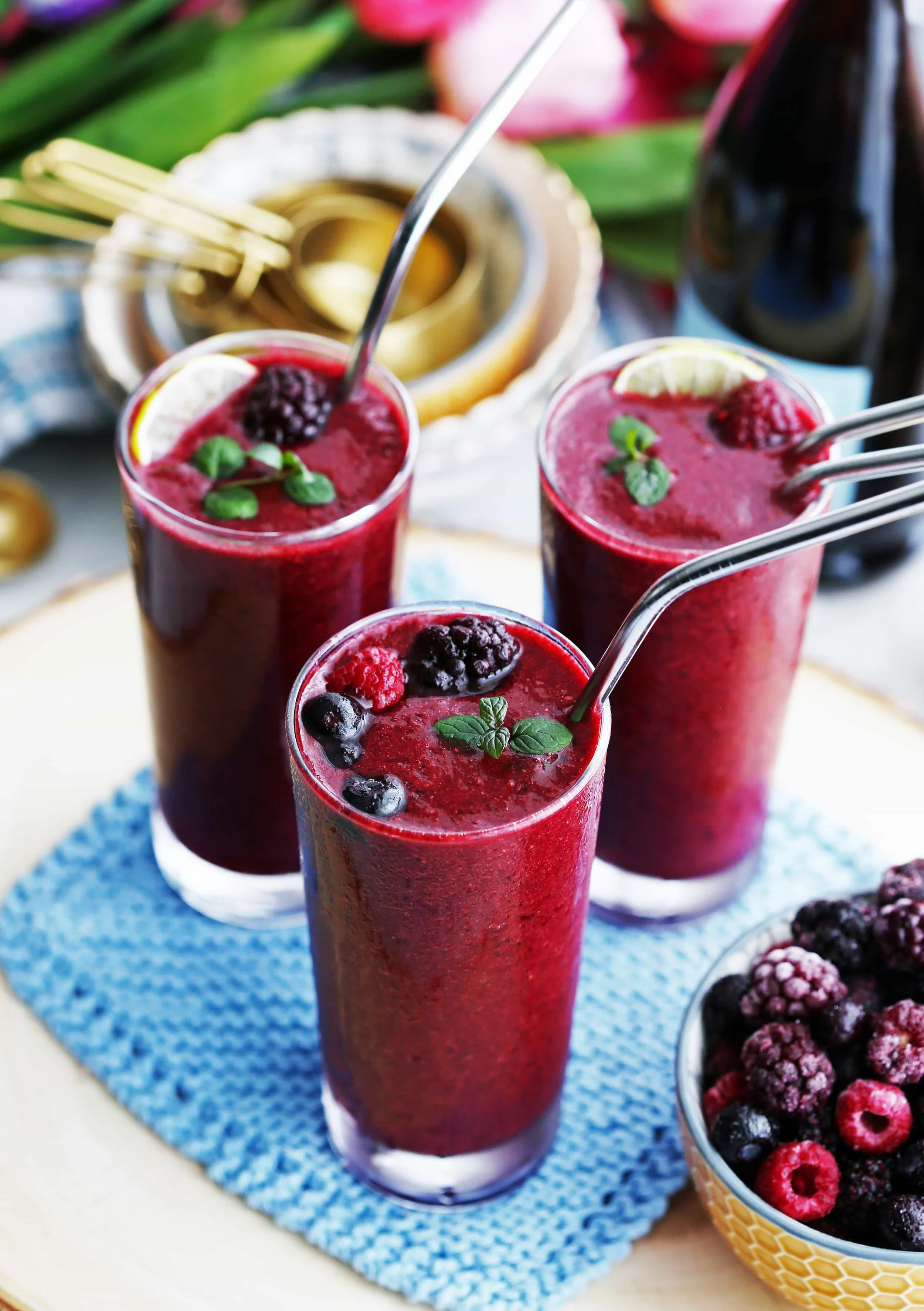 Three glass tumblers full of Mixed Berry Prosecco Wine Slushy cocktail with straws, mint, lime, and berry garnish.