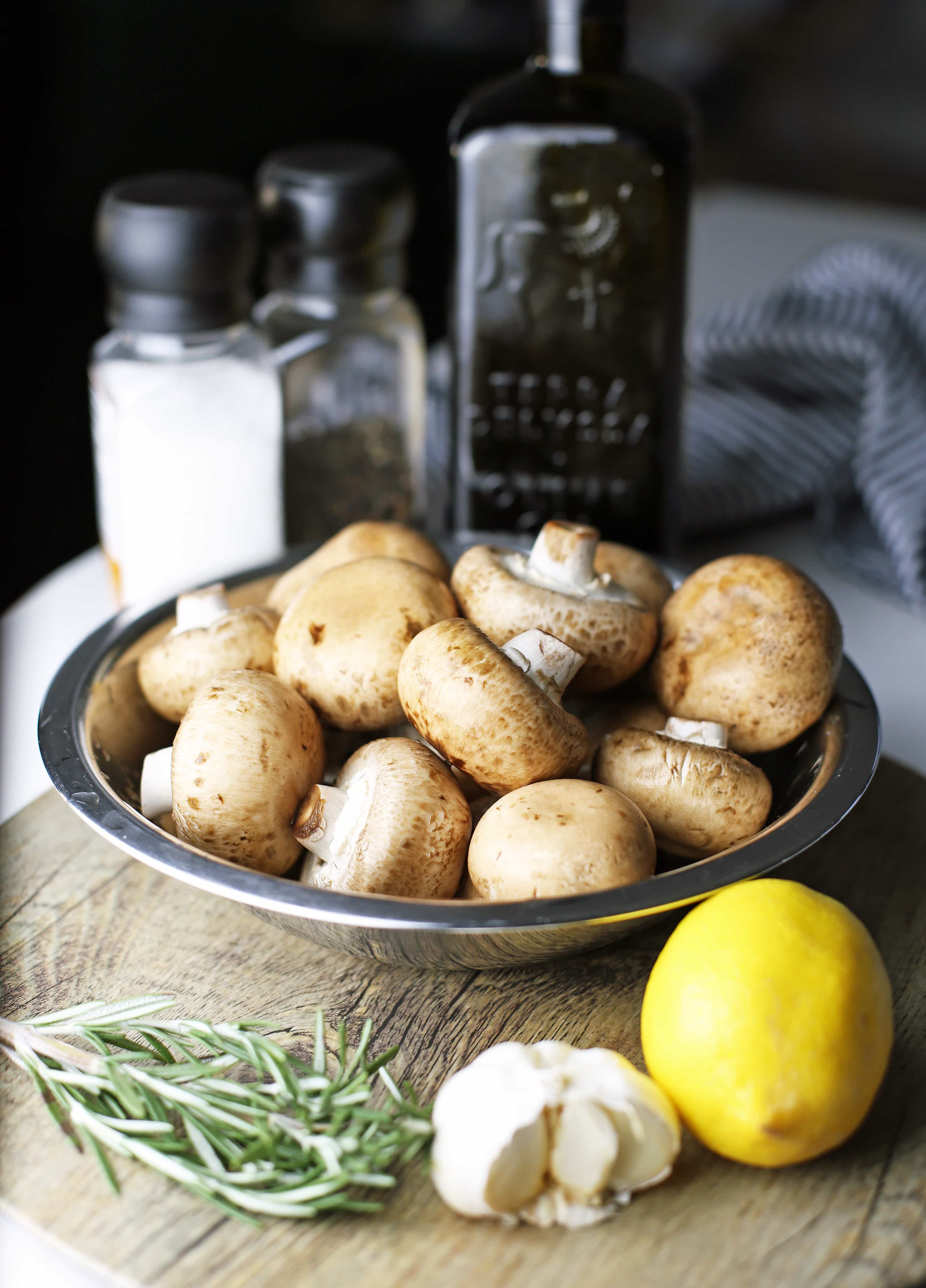 A bowl of fresh cremini mushrooms surrounded by fresh rosemary, garlic, and lemon.