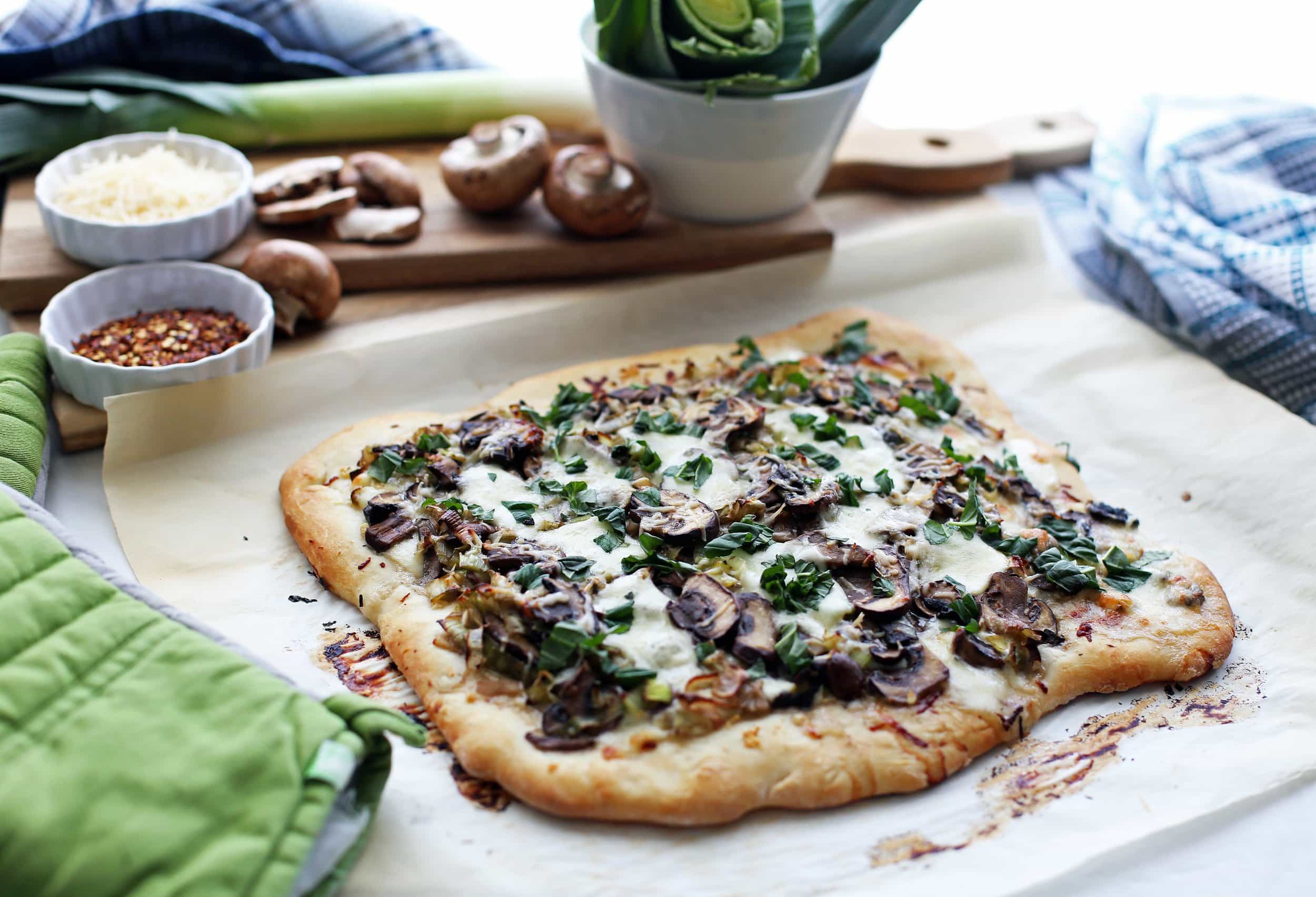 Side view of leek mushroom mozzarella pizza with fresh mushrooms and leeks in the background.