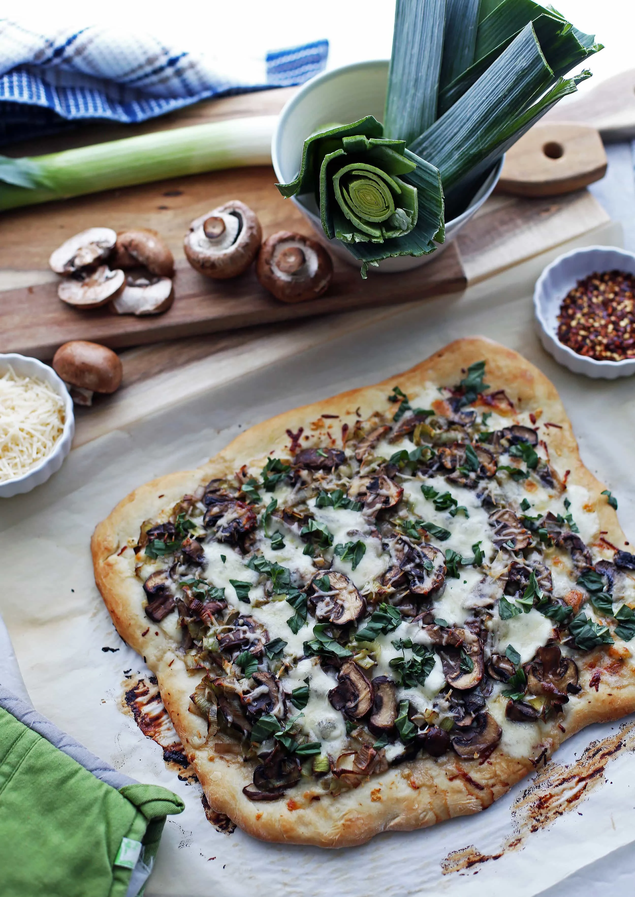 Overhead view of of leek mushroom mozzarella pizza with fresh mushrooms, leeks, and cheese in the background.