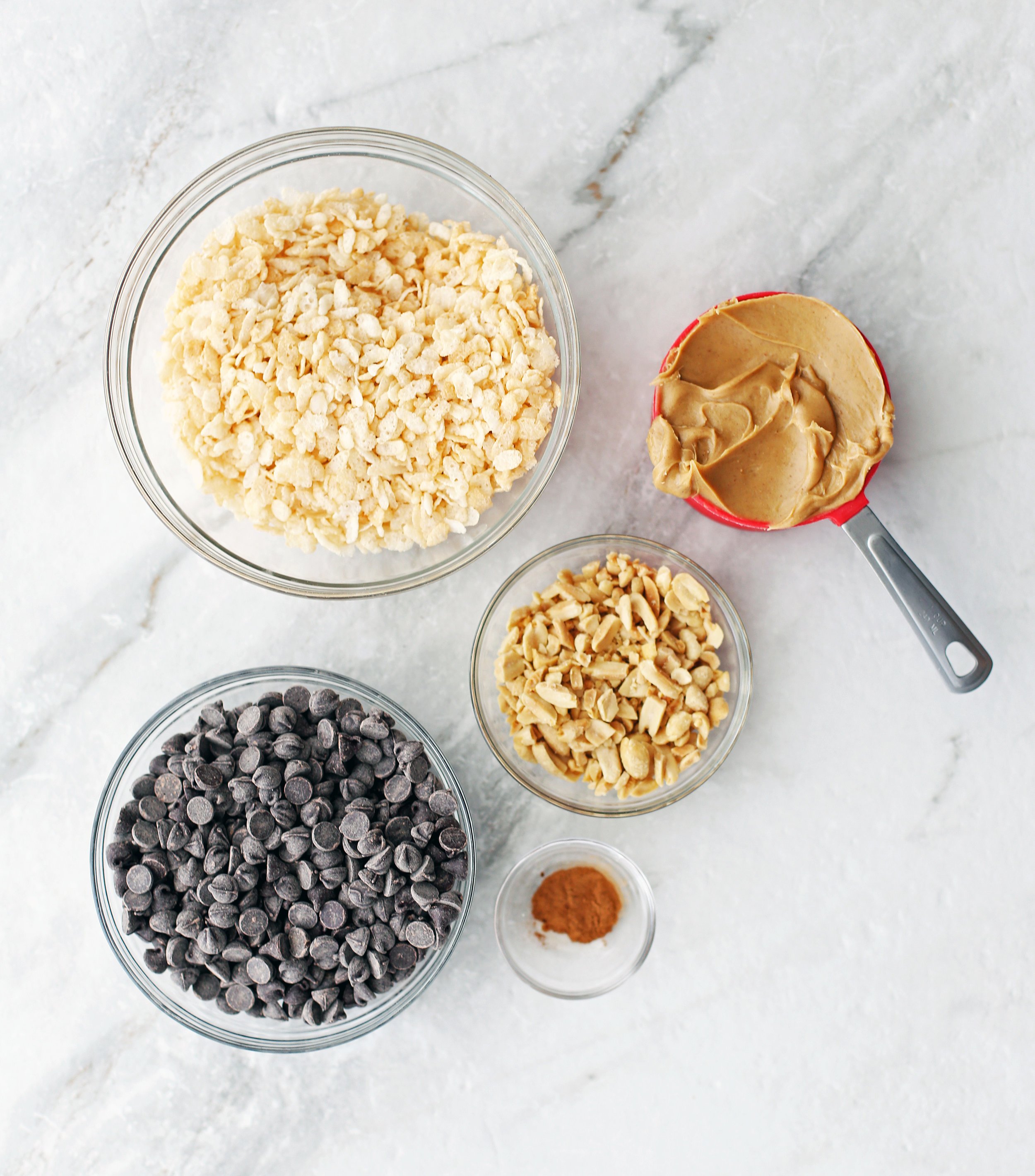 Bowls of chocolate chips, chopped peanuts, peanut butter, rice cereal, and ground cinnamon.