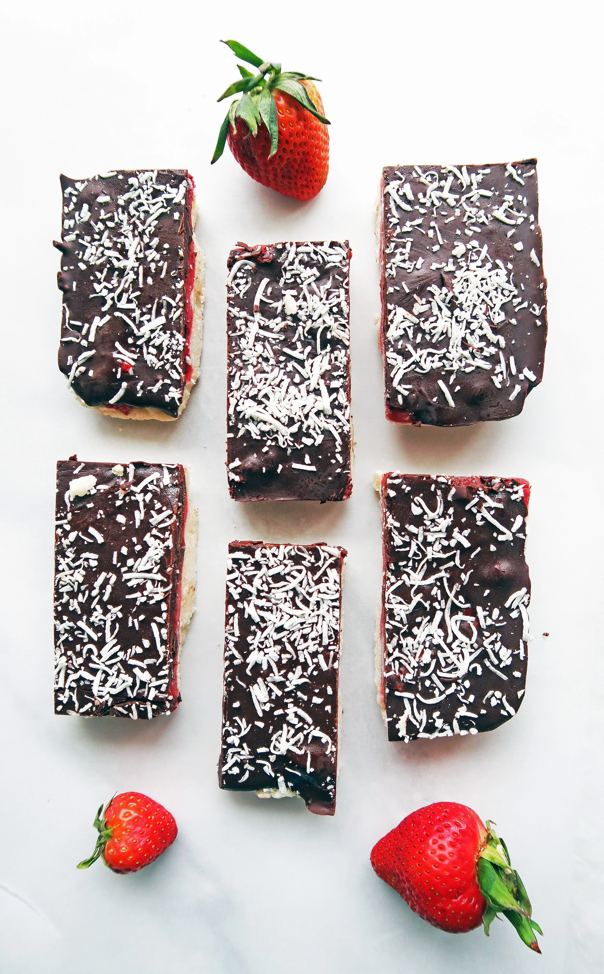No-Bake, vegan, and gluten-free Chocolate Strawberry Coconut Bars on parchment paper with strawberries around them.  