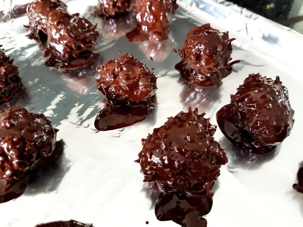 Coconut balls freshly coated with rich chocolate spread out on a baking sheet.