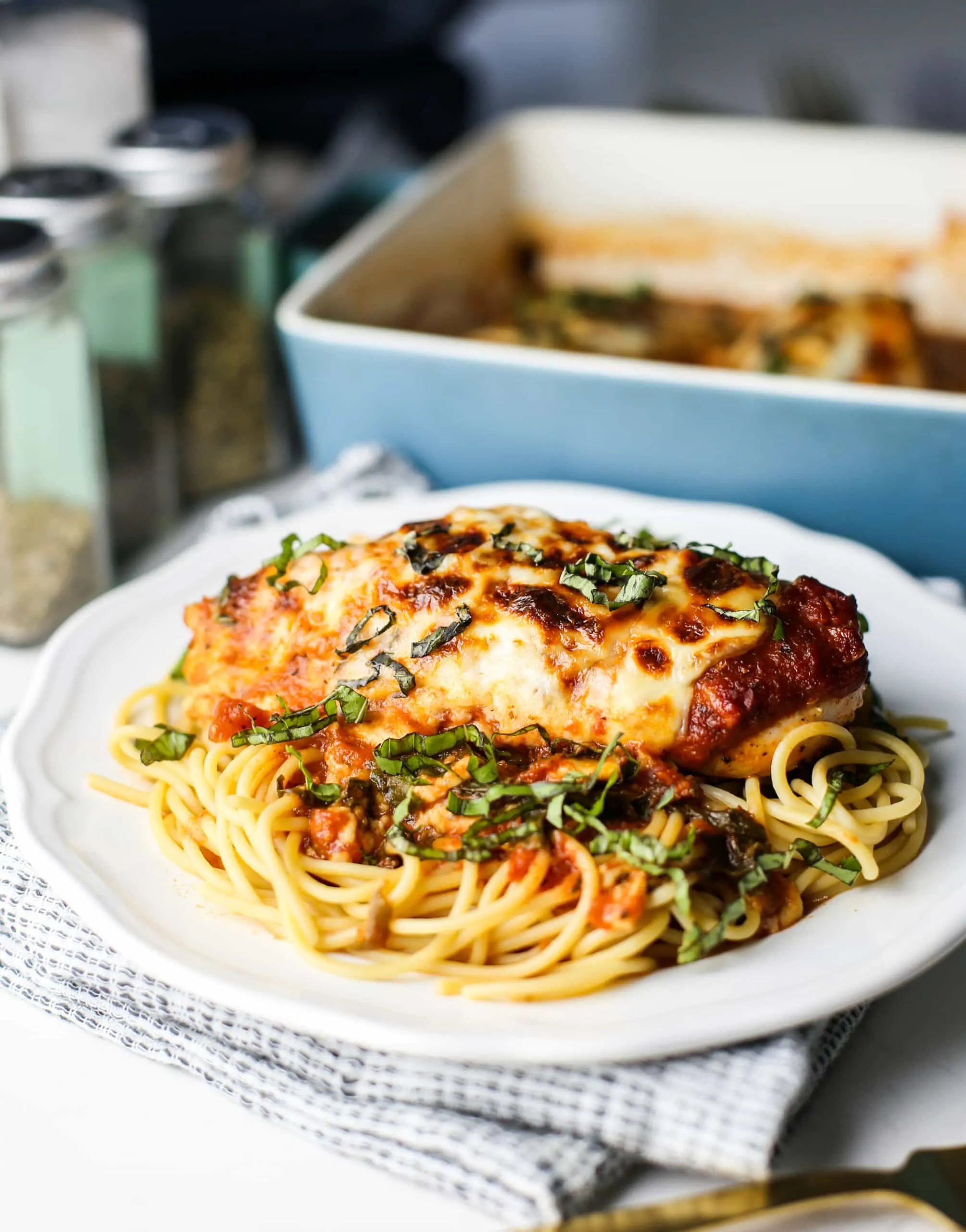 A side view of baked chicken with mozzeralla and marinara sauce on top of spaghetti in a white plate.