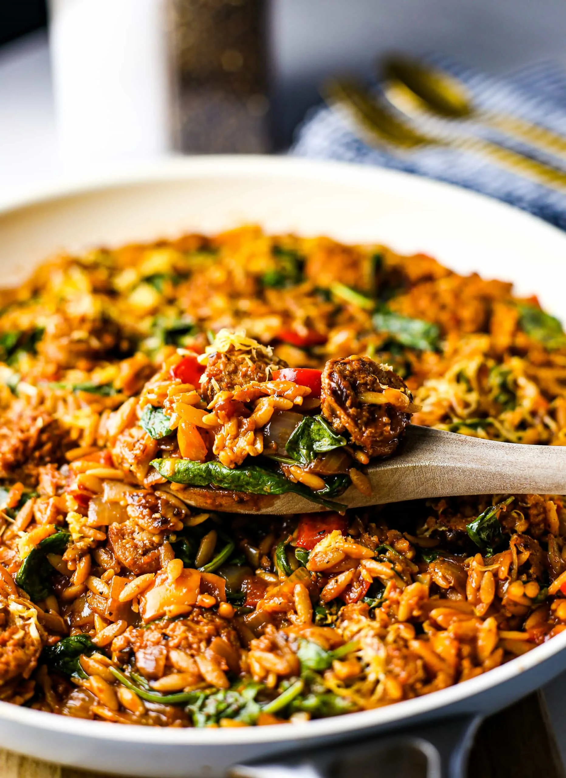 A closeup view of a wooden spoon holding a scoop of Italian sausage, vegetables, orzo pasta on top of more pasta in a frying pan.