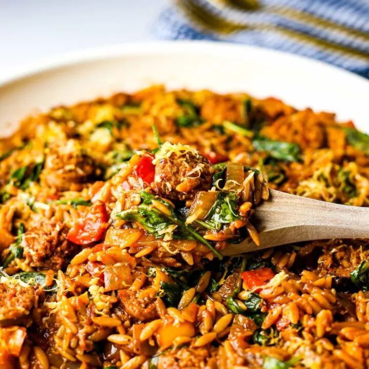 A closeup view of a wooden spoon holding a scoop of Italian sausage and orzo pasta on top of more pasta in a frying pan.