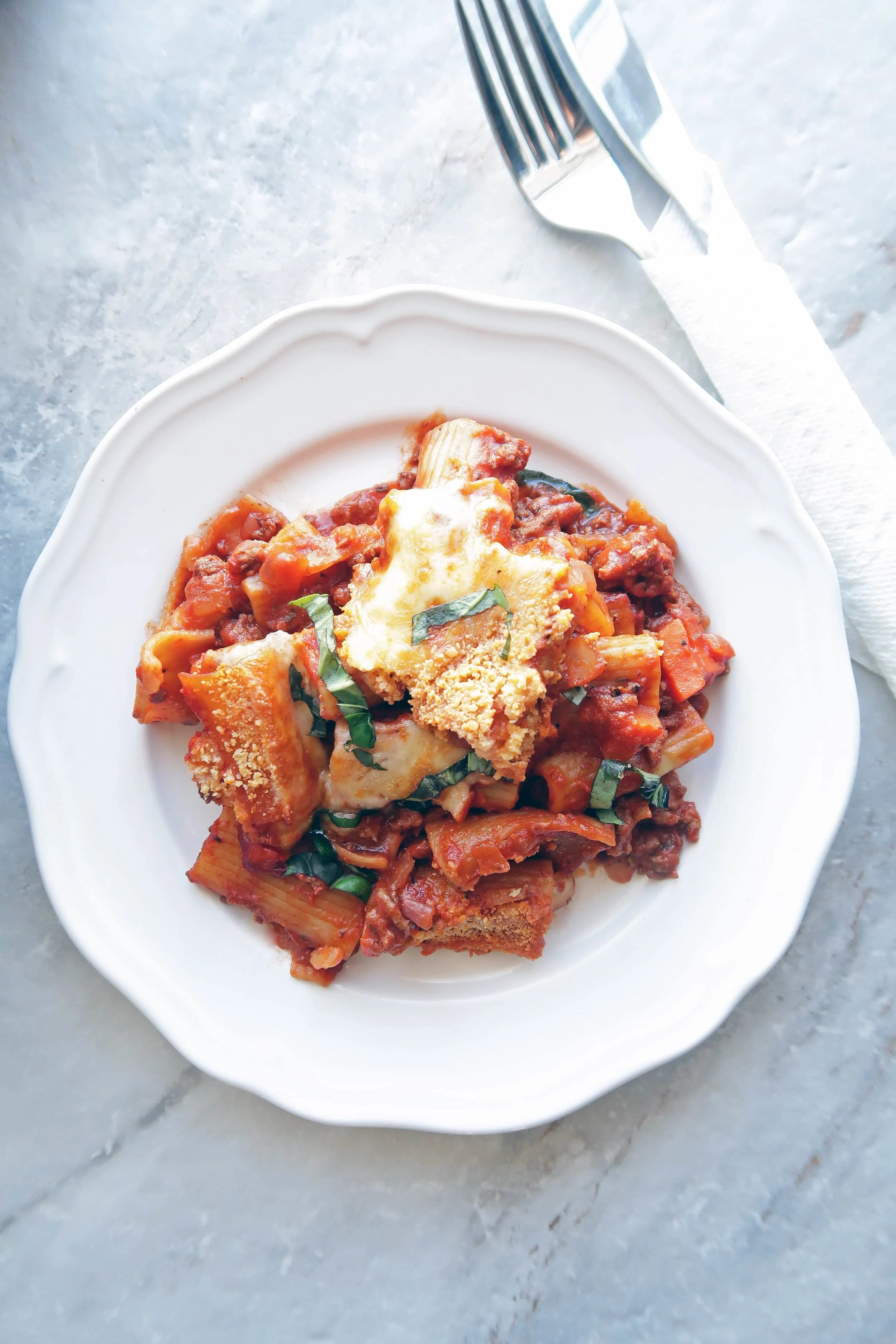 A heaping scoop of One-Pot Beef Pasta Casserole on a white plate with fork and knife to its side.