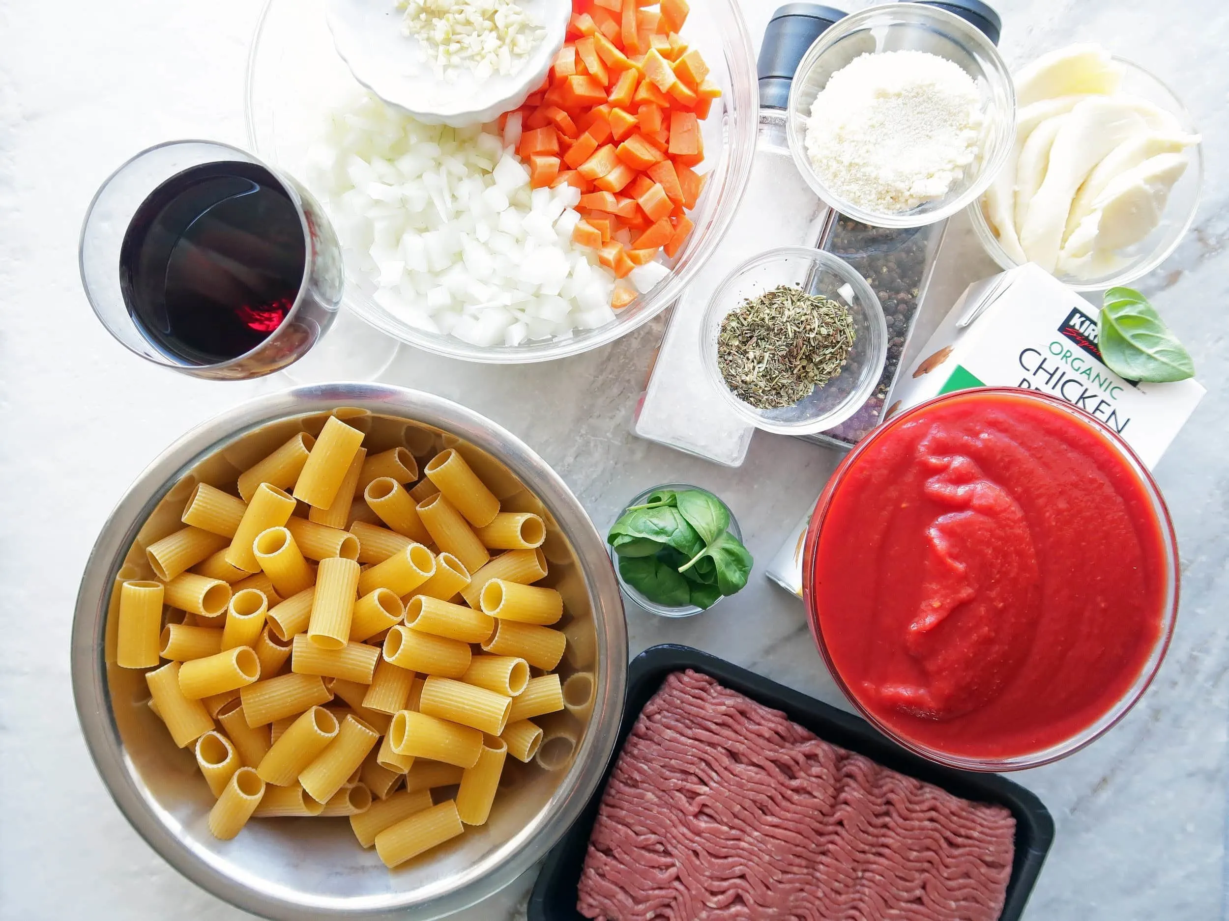 Bowls of dry rigatoni, chopped onions and peppers, cheese, crushed tomatoes, and herbs next to ground beef.