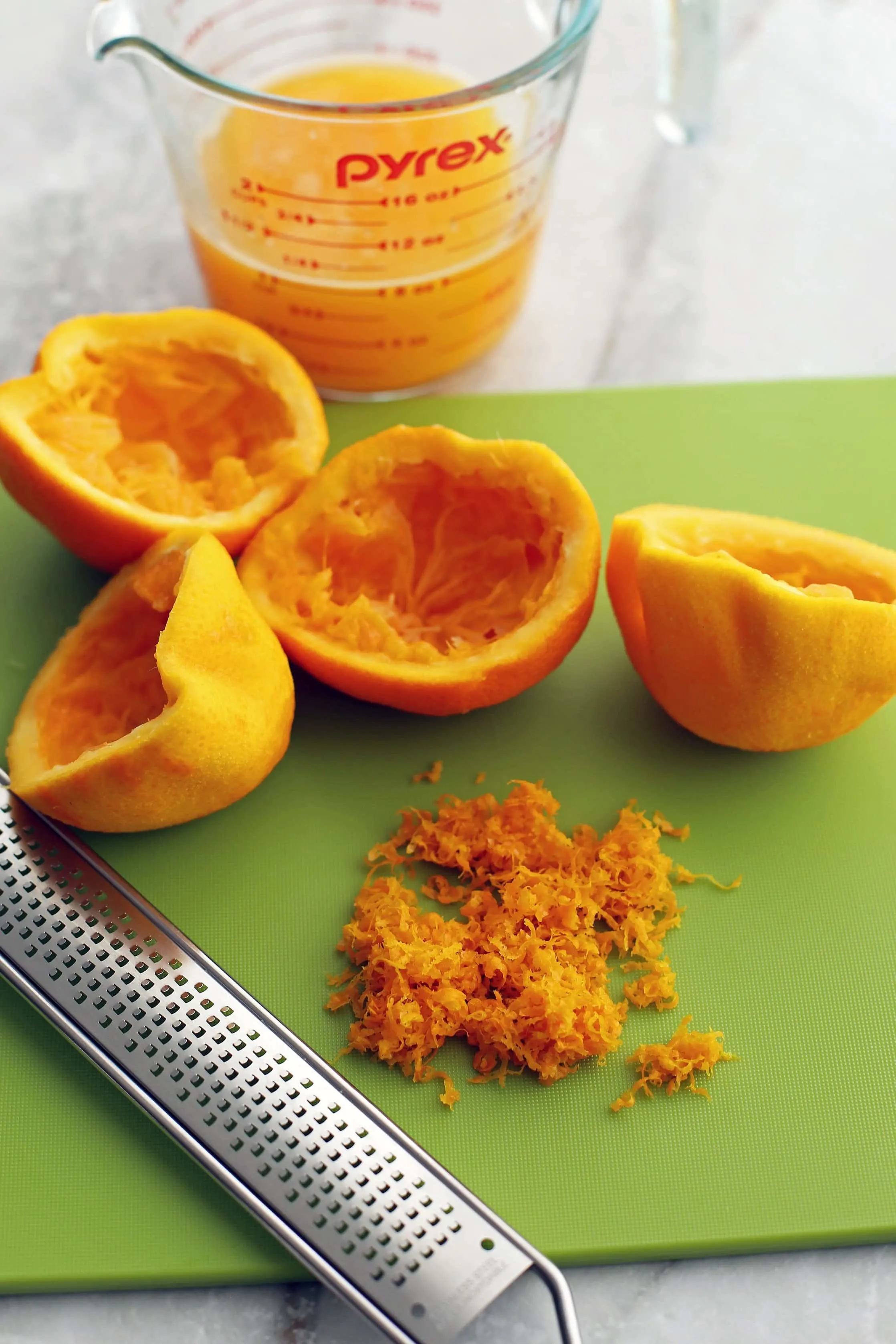 Orange zest and juiced oranges on a green cutting board and a glass measuring cup filled with orange juice.