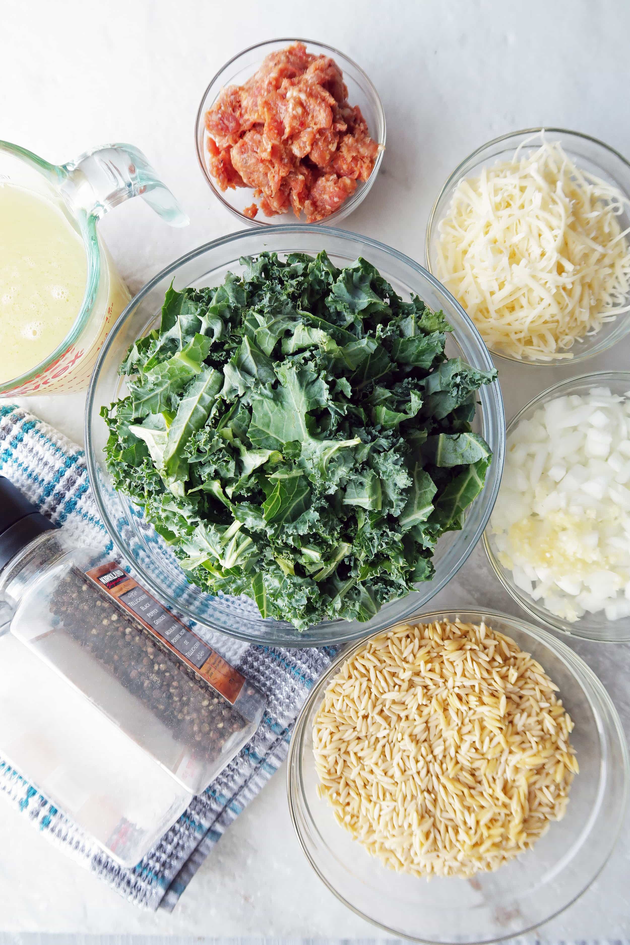 Orzo, onions, kale, cheese, sausage, chicken broth in different glass bowls with salt and pepper grinders.