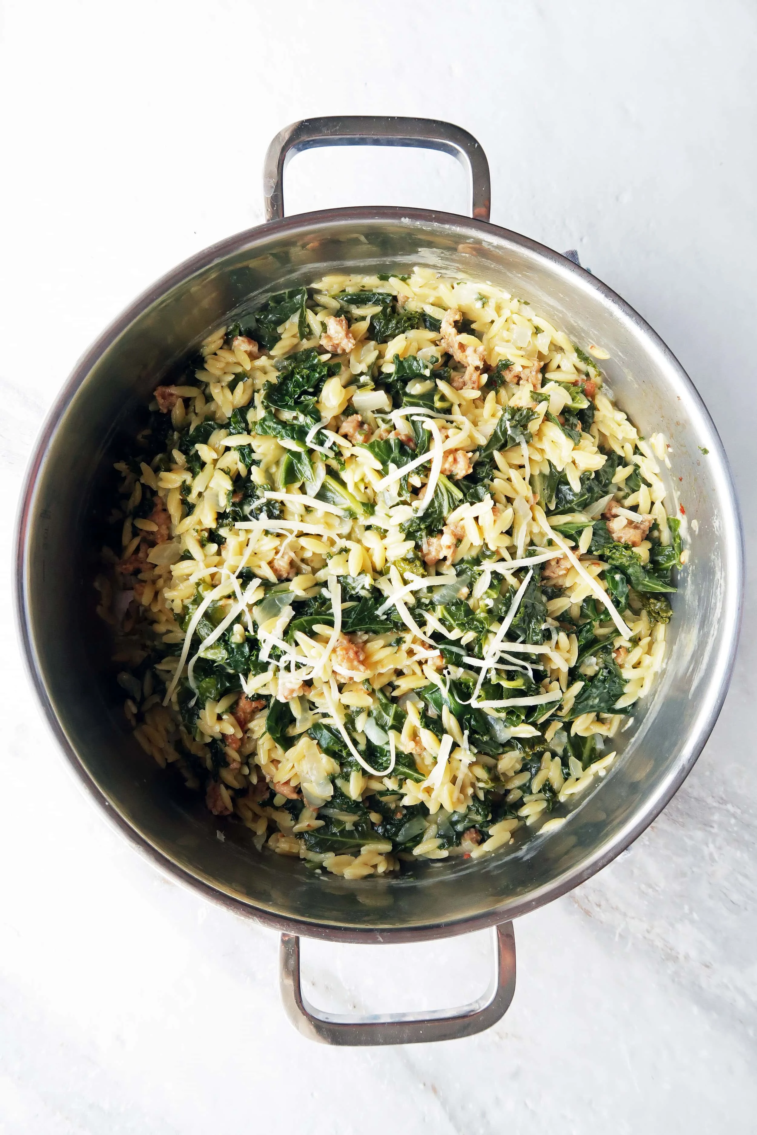 Orzo pasta with Italian sausage and kale with shredded cheese on top in a metal pot.