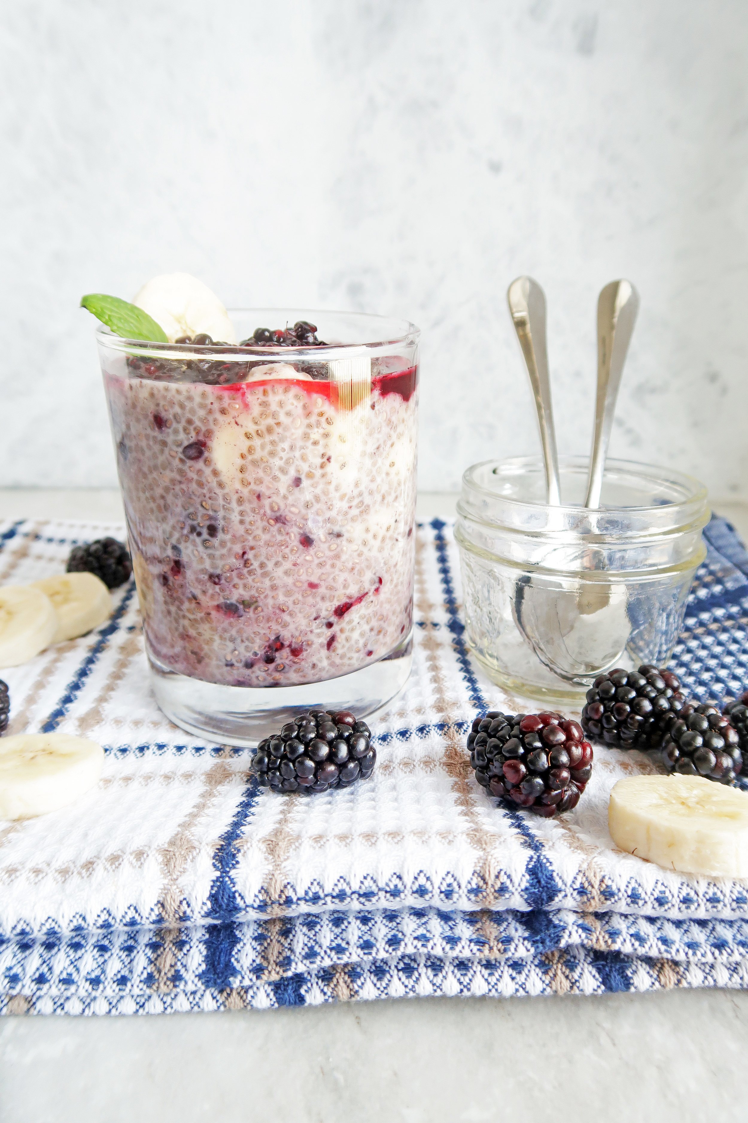 Overnight Chia Seed Pudding with Blackberries and Bananas in glass tumbler along with spoons in a small glass jar and blackberries and banana slices around it.