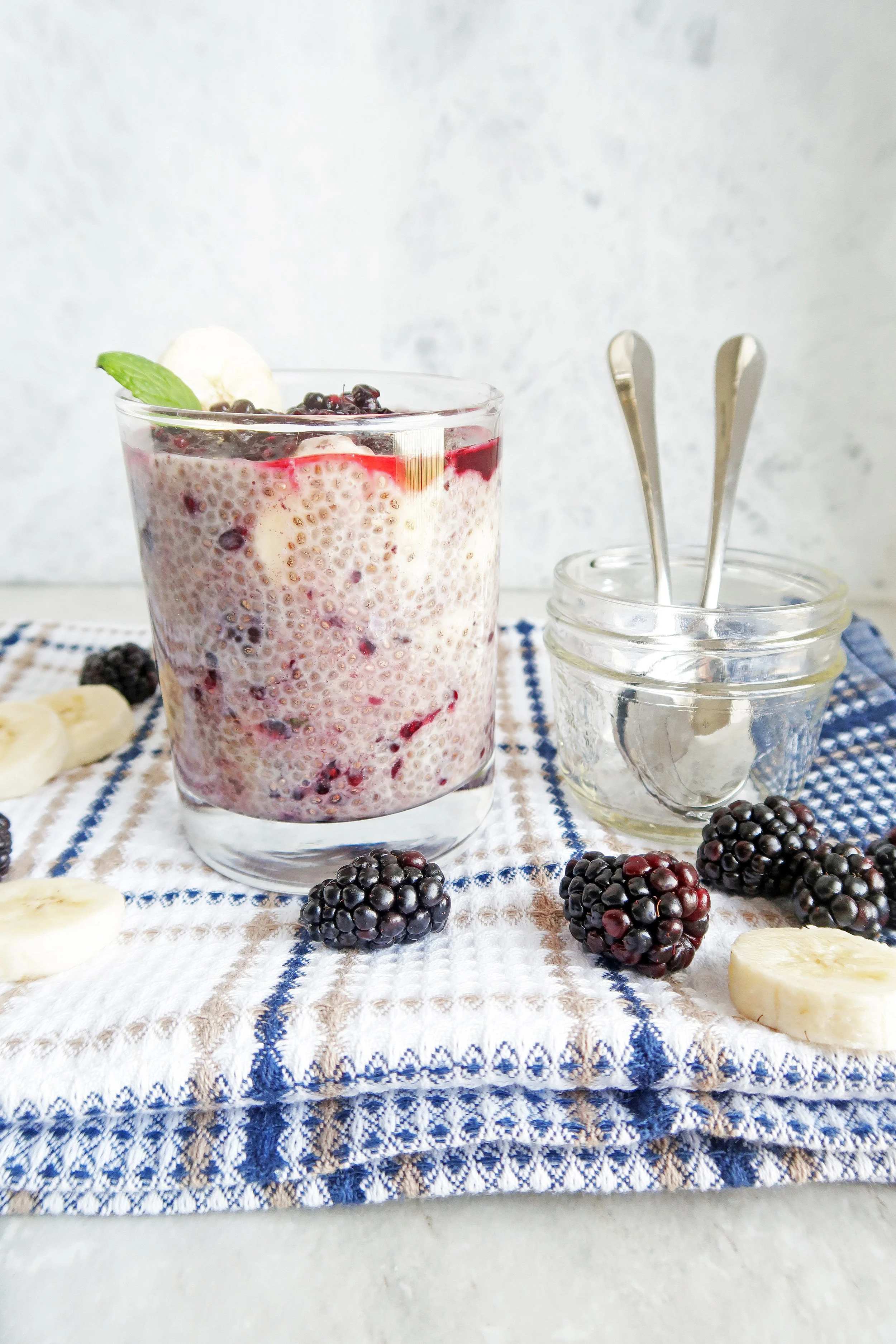 Overnight Chia Seed Pudding with Blackberries and Bananas in glass tumbler along with spoons in a small glass jar and blackberries and banana slices around it.