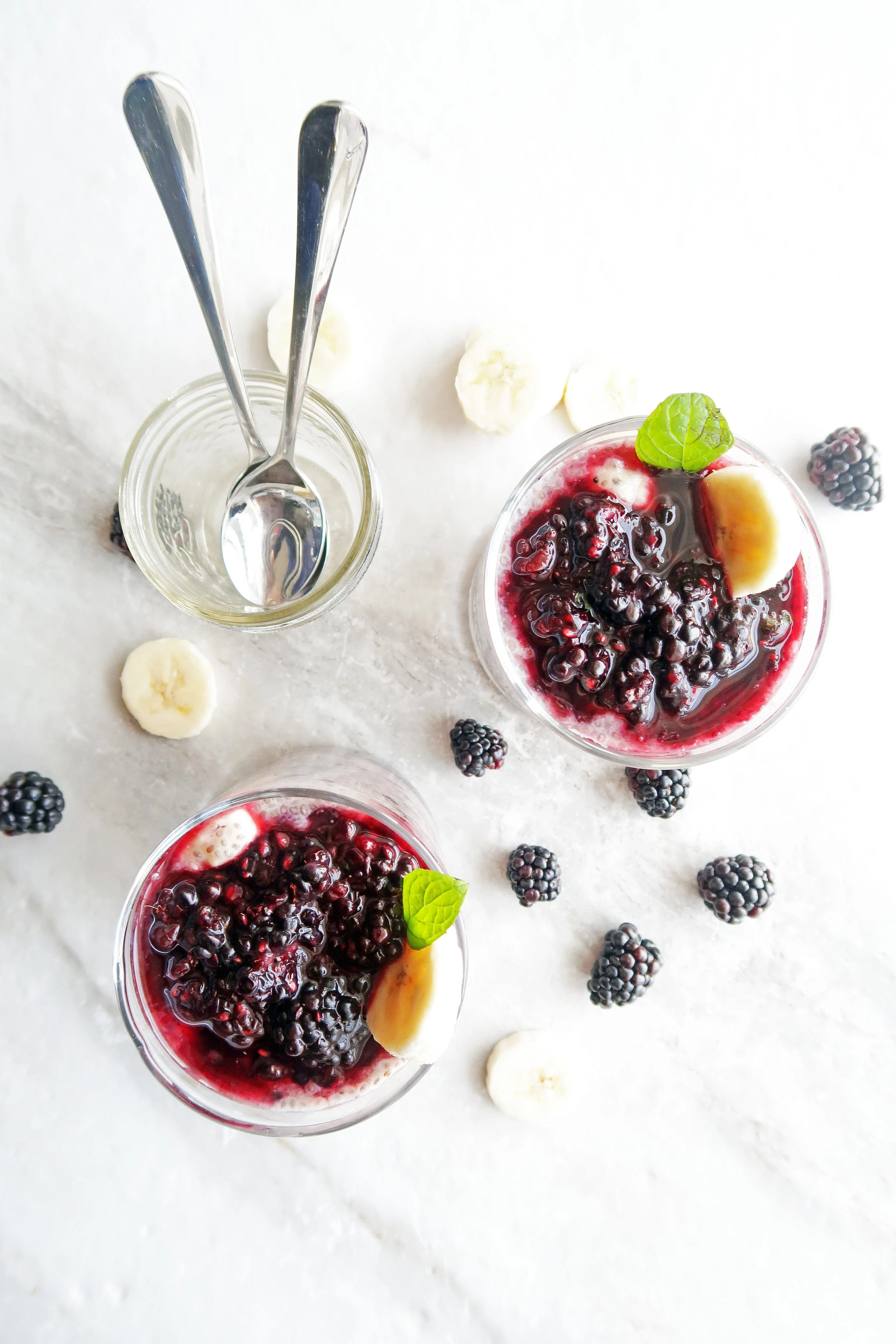 Overnight Chia Seed Pudding with Blackberries and Bananas in two glass tumblers along with spoons in a small glass jar and blackberries and banana slices around it.