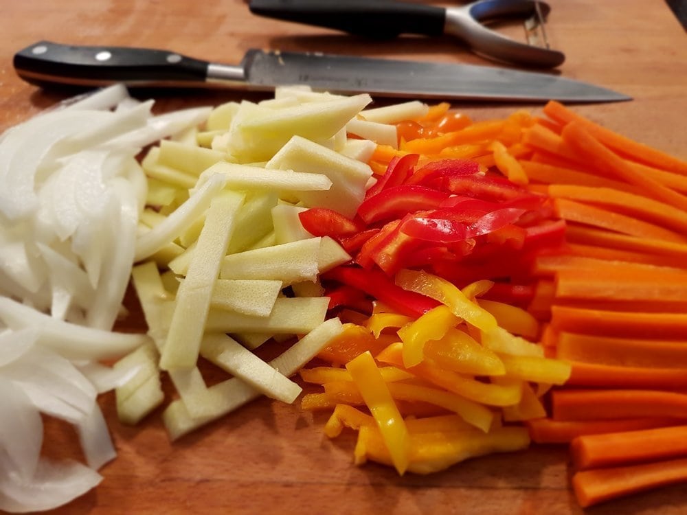 Thin strips of onions, zucchini, peppers, and carrots neatly aligned on a cutting board.