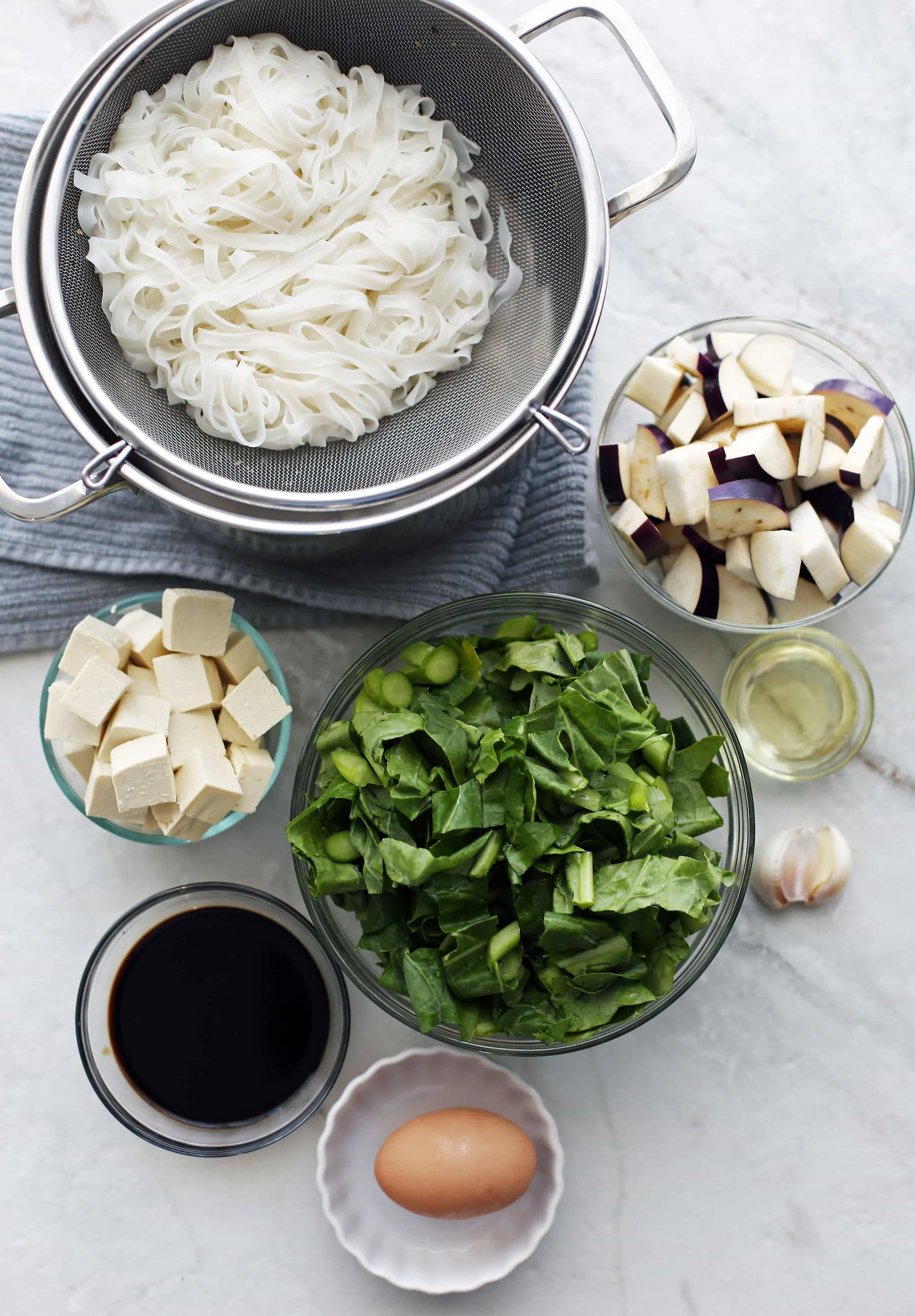Bowls of cooked rice noodles, tofu, gai lan, eggplants, egg, soy sauce, and garlic.